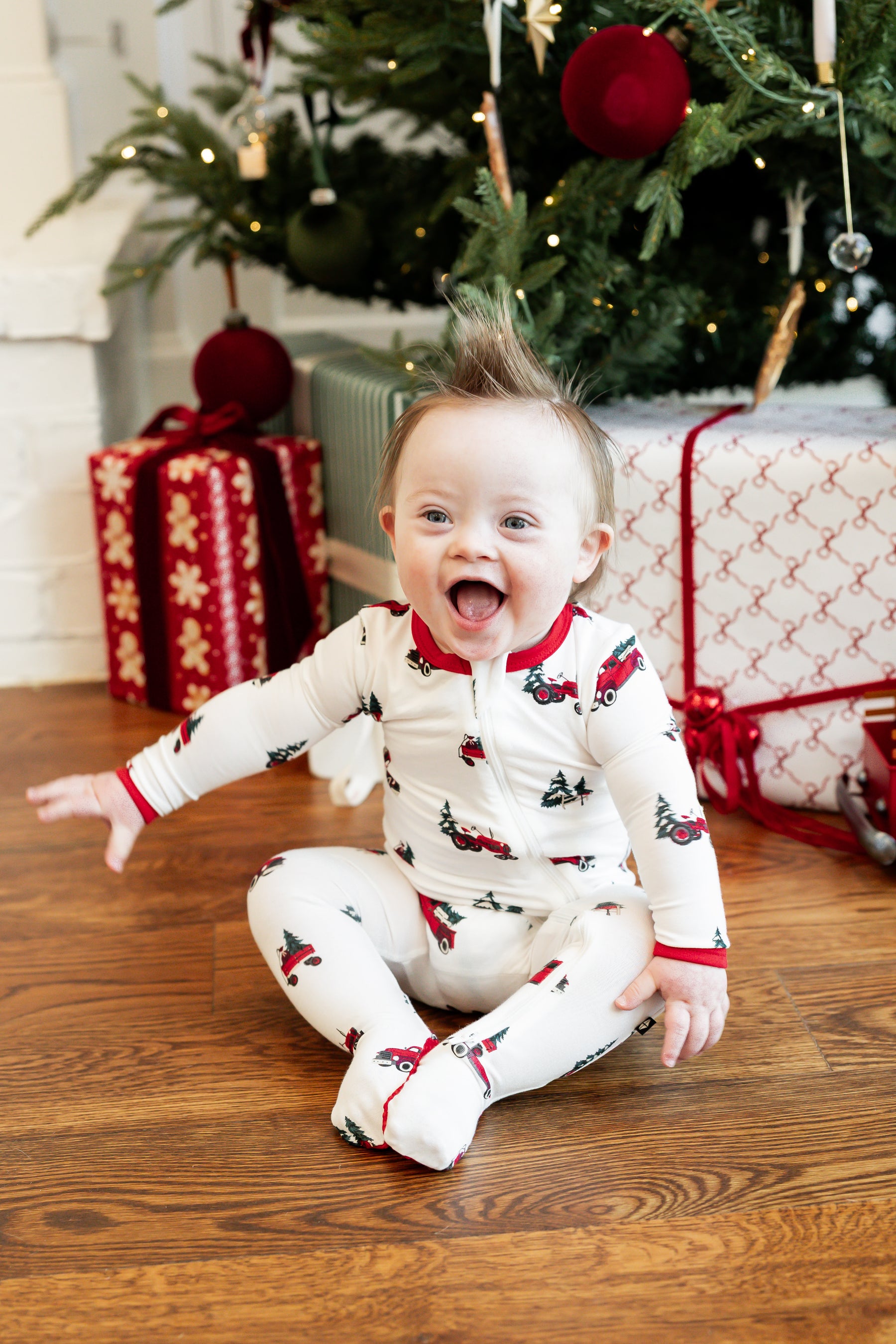 A baby sitting on a wooden floor in front of a Christmas tree, wearing a white footed zip-up pajama romper with a red and green holiday print. Gift-wrapped presents and the decorated tree are visible in the background.