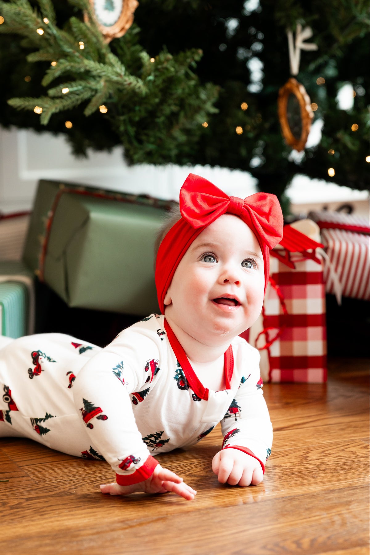 A baby wearing a white Christmas-themed romper with red trim and a matching red bow headband lies on a wooden floor under a decorated Christmas tree with wrapped presents.