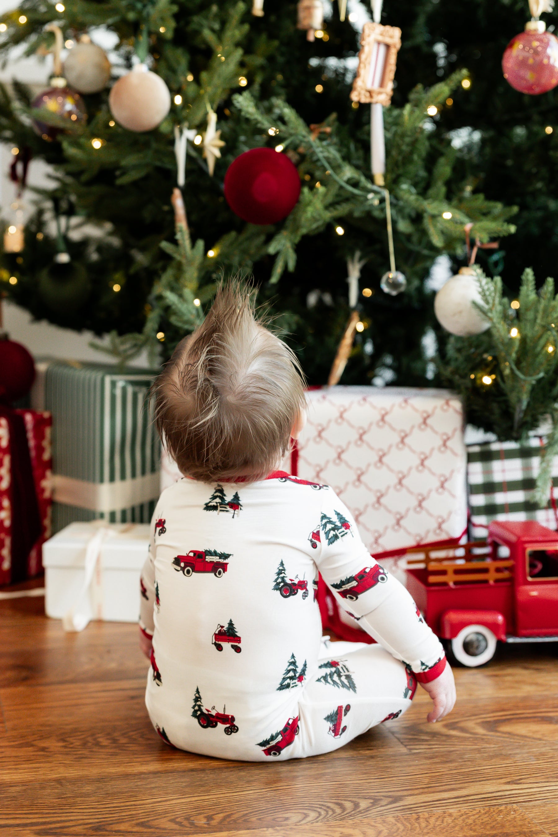 A toddler sits on a wooden floor in front of a decorated Christmas tree, wearing a white long-sleeve pajama top with a red and green truck and tree print. Gifts and a red toy truck are visible around the tree.