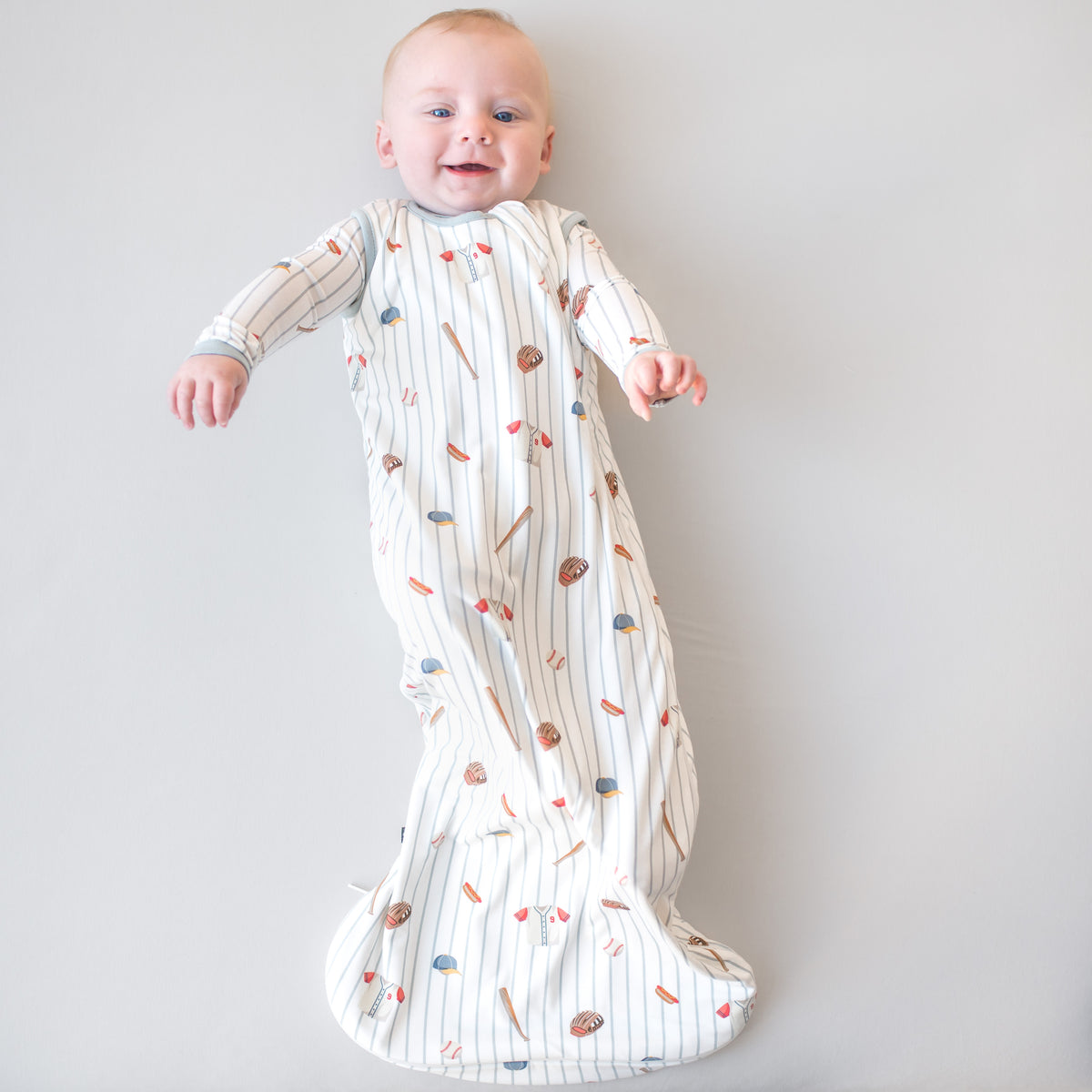 Infant laying on a light neutral surface wearing the Sleep Bag in Home Run 0.5 with matching romper on underneath
