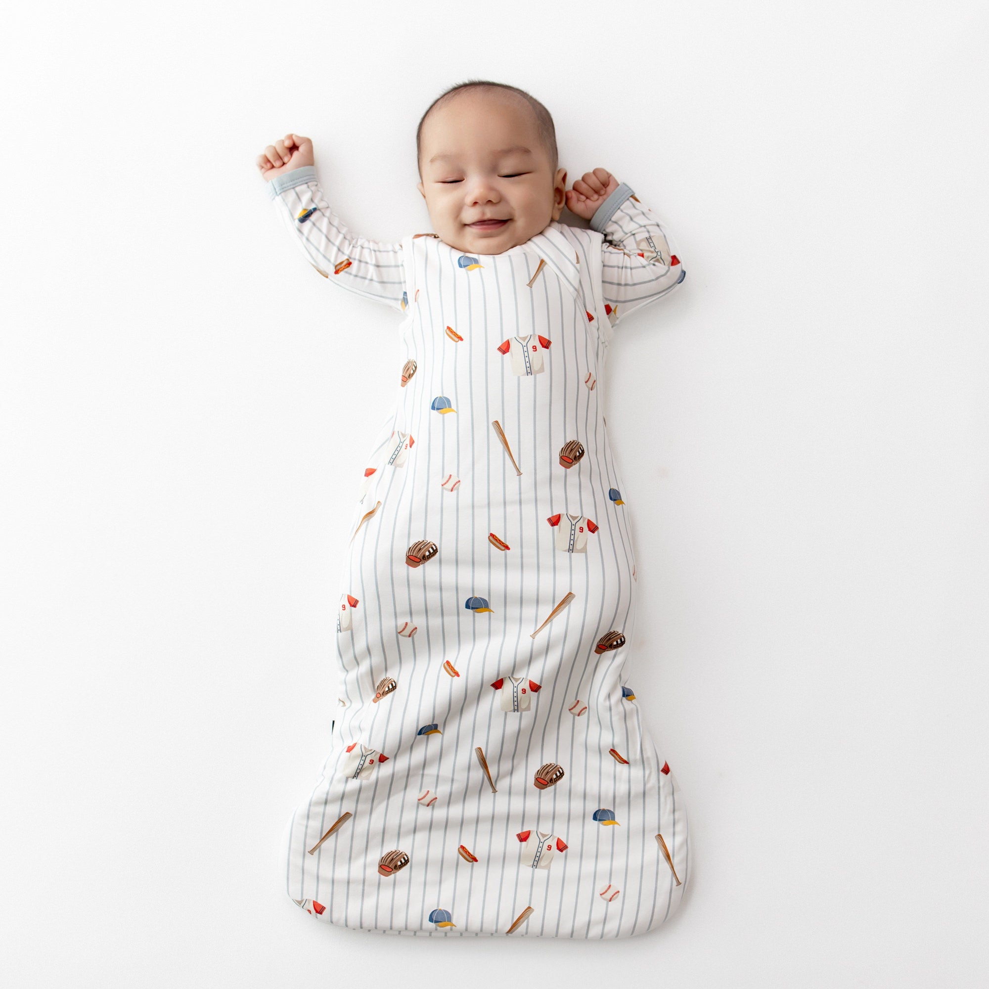 Baby stretching out on a white surface wearing the Sleep Bag in Home Run 1.0 with a matching romper
