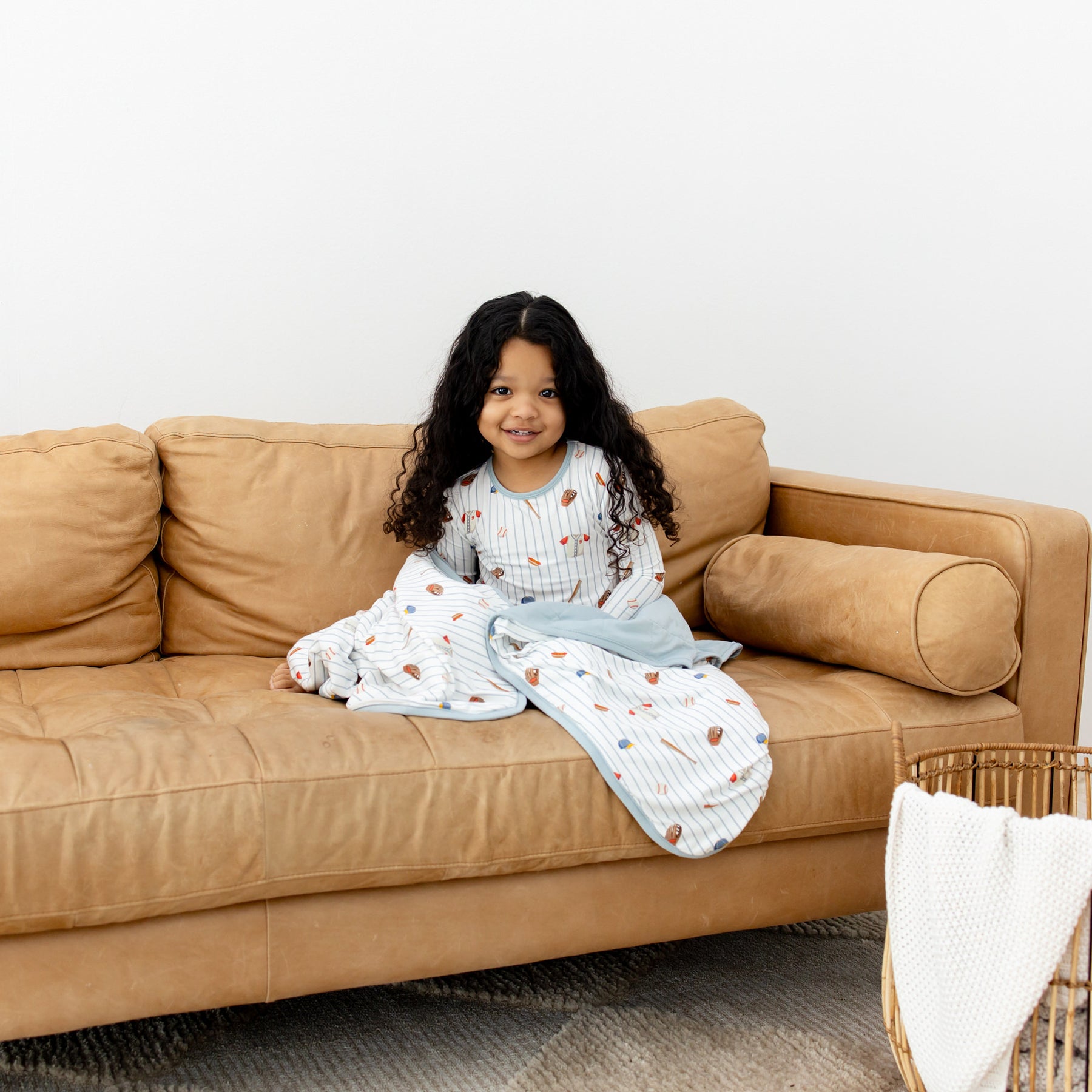 Young girl sitting on a brown couch with the Toddler Blanket in Home Run 1.0 around her wearing matching long sleeve pajamas