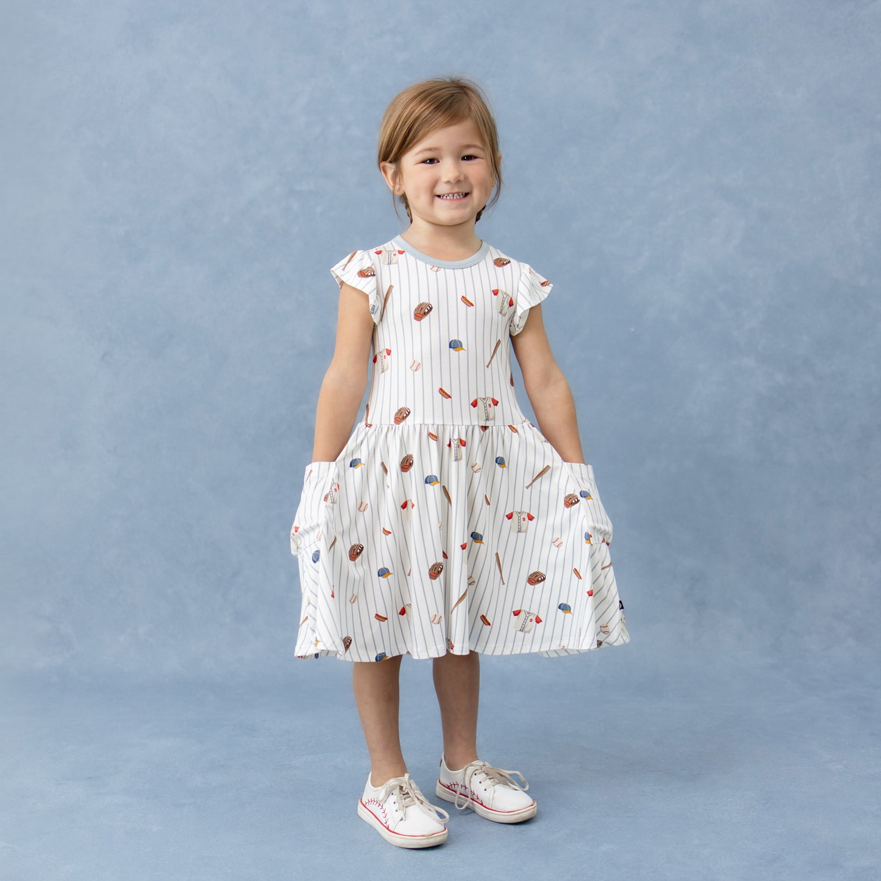 Young girl standing in front of a marbled blue background wearing the Pocket Dress in Home Run with her hands in the pockets