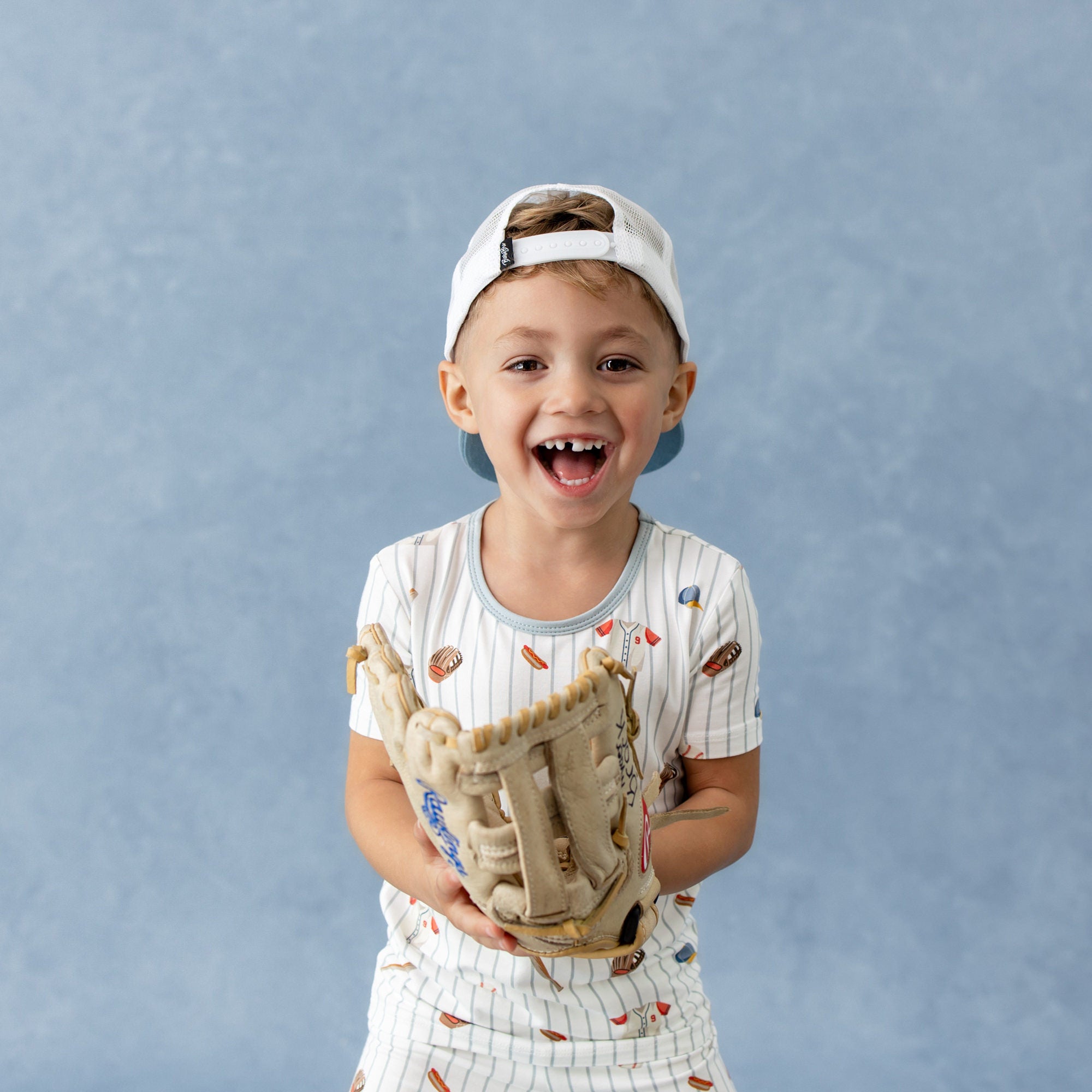 Close up of young smiling boy wearing the Short Sleeve Pajamas in Home Run and baseball glove with a backwards baseball cap