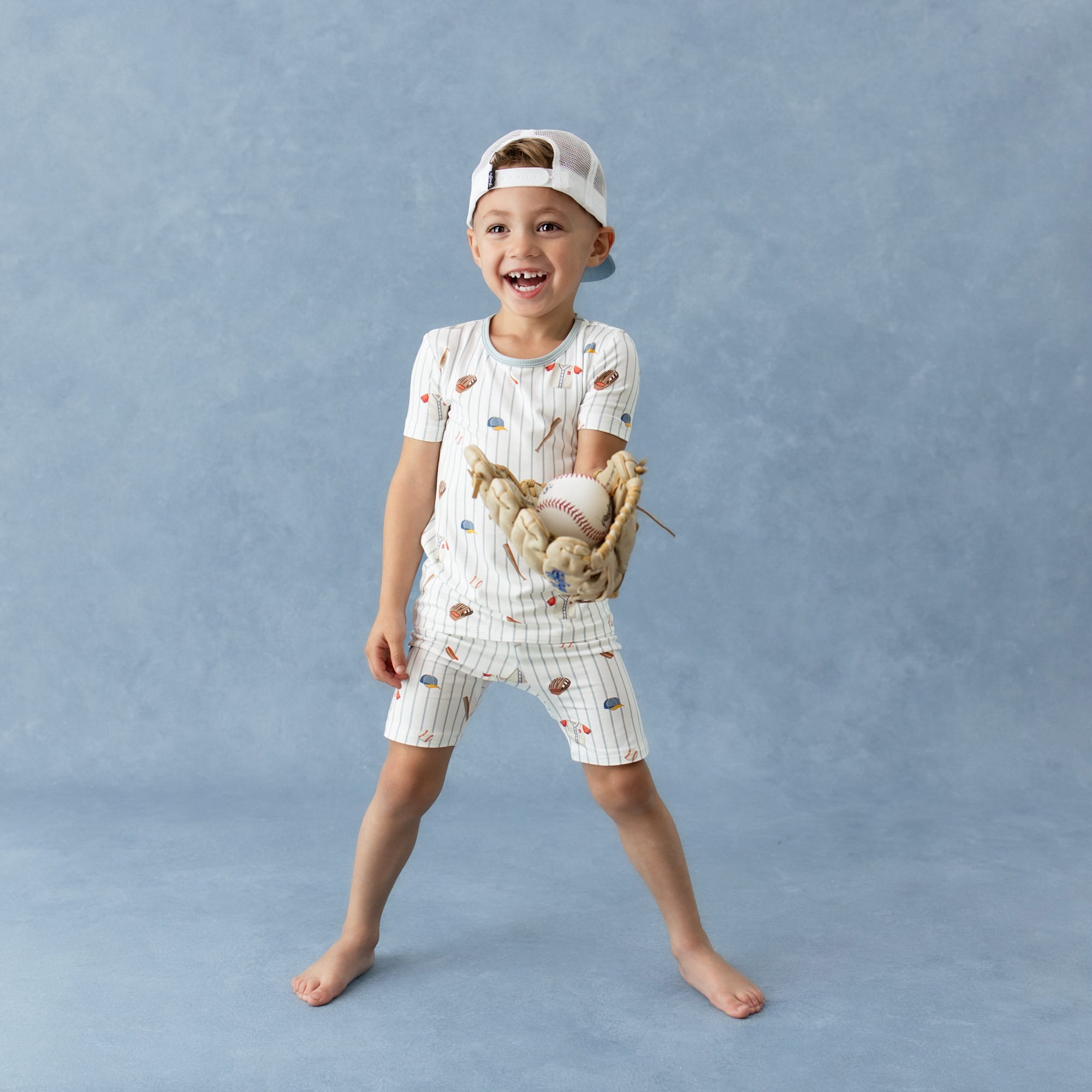 Young smiling boy wearing the Short Sleeve Pajamas in Home Run and backwards baseball cap with a baseball glove on his hand holding a baseball