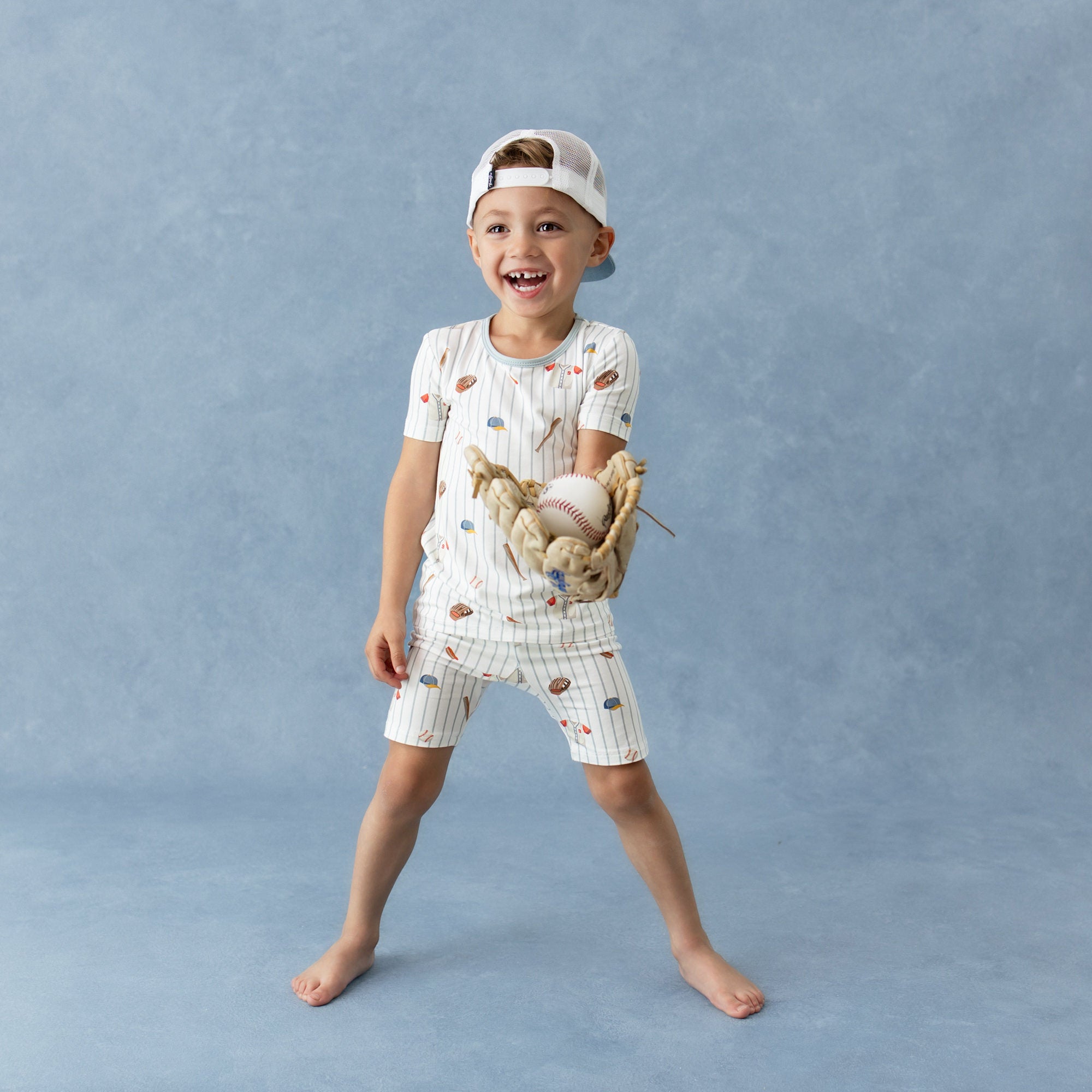 Young smiling boy wearing the Short Sleeve Pajamas in Home Run and backwards baseball cap with a baseball glove on his hand holding a baseball