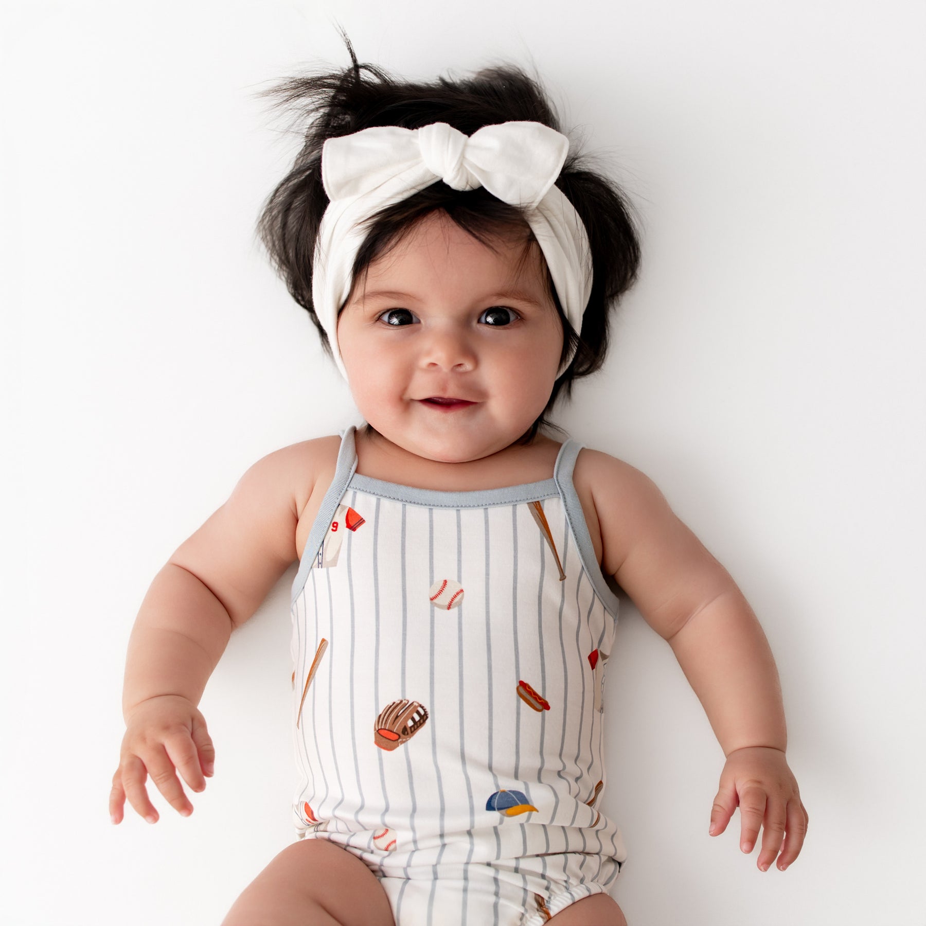 Young infant wearing the Spaghetti Strap Leotard in Home Run paired with a white knotted bow headband