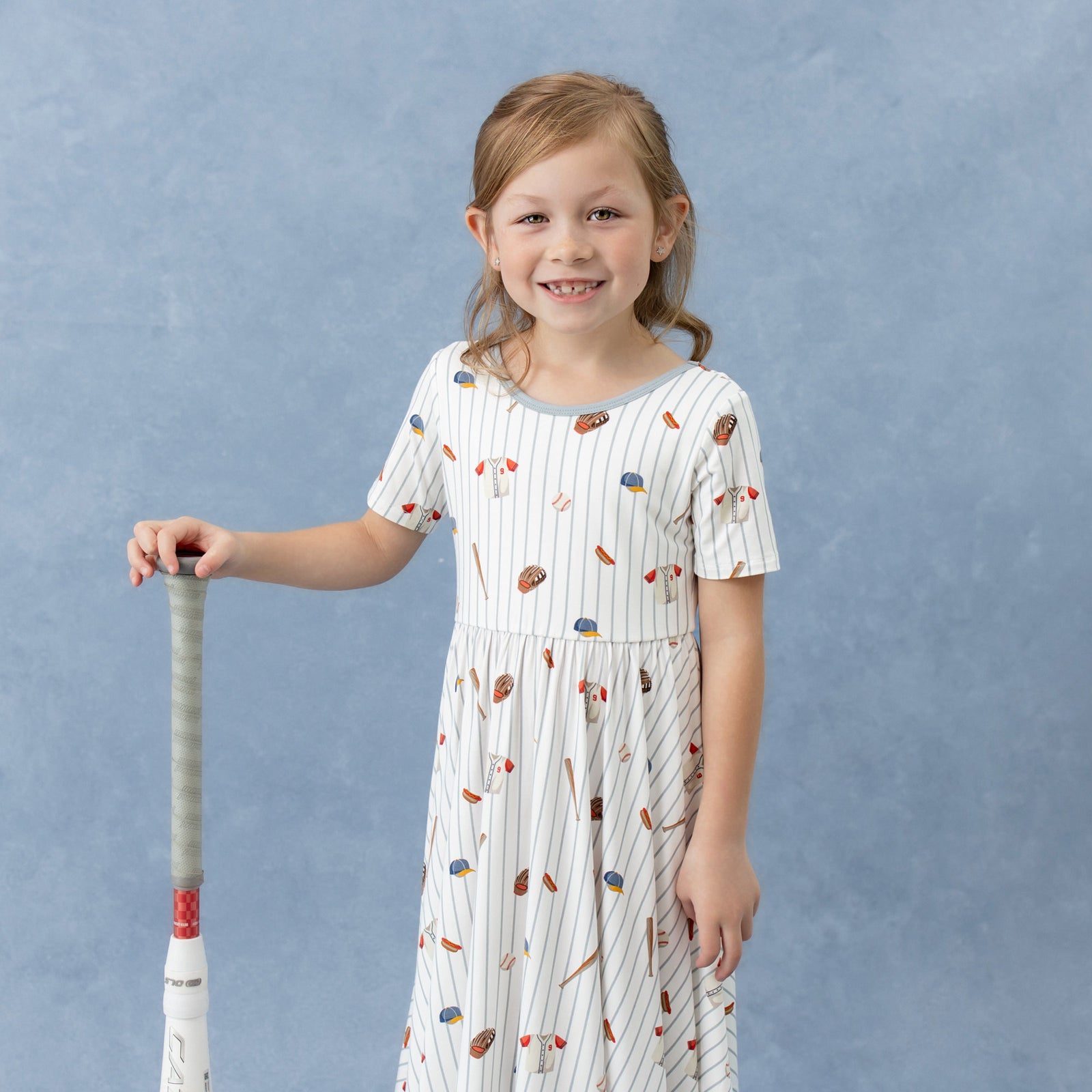 Close up of young girl wearing the Twirl Dress in Home Run standing beside a baseball bat
