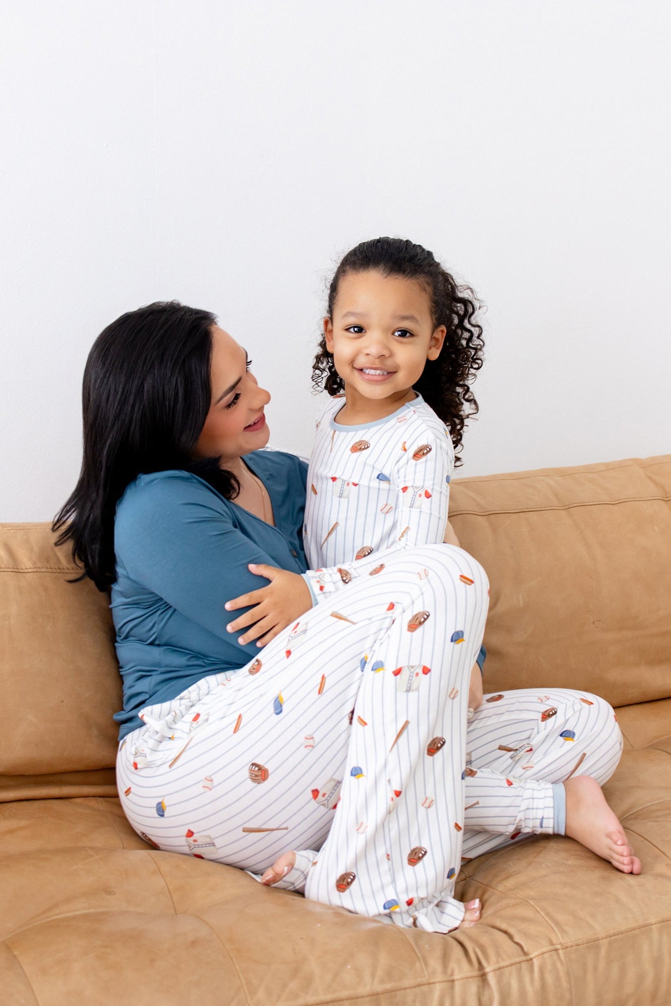 Female sitting on a couch wearing the Women's Lounge Pants in Home Run with her daughter who is matching in long sleeve pajamas
