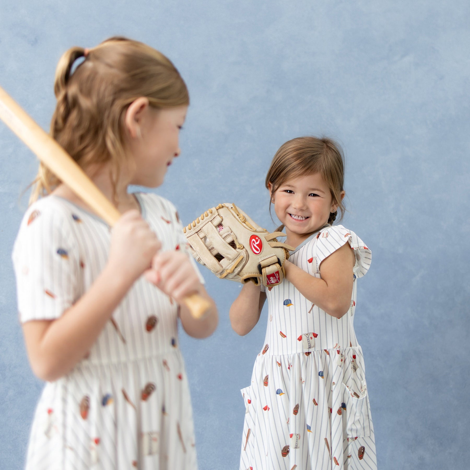Two sisters standing in front of one another both wearing Home Run dresses holding baseball accessories