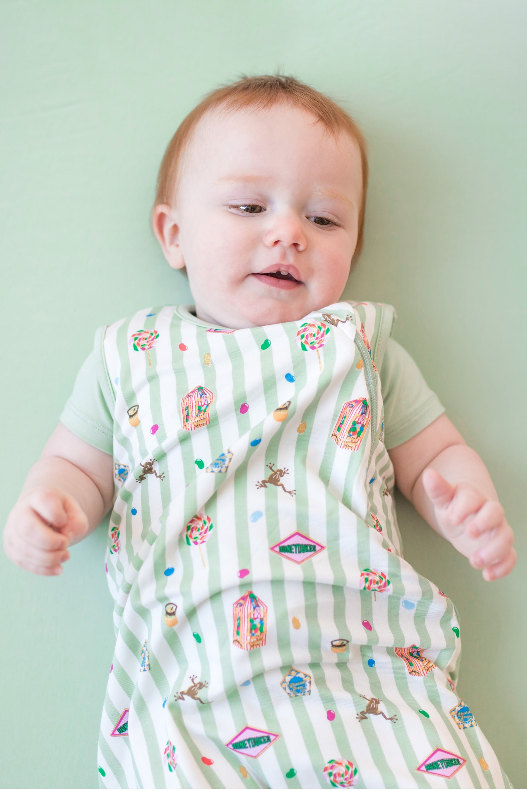 Baby wearing a green and white striped sleep bag with colorful Harry Potter patterns on a green background