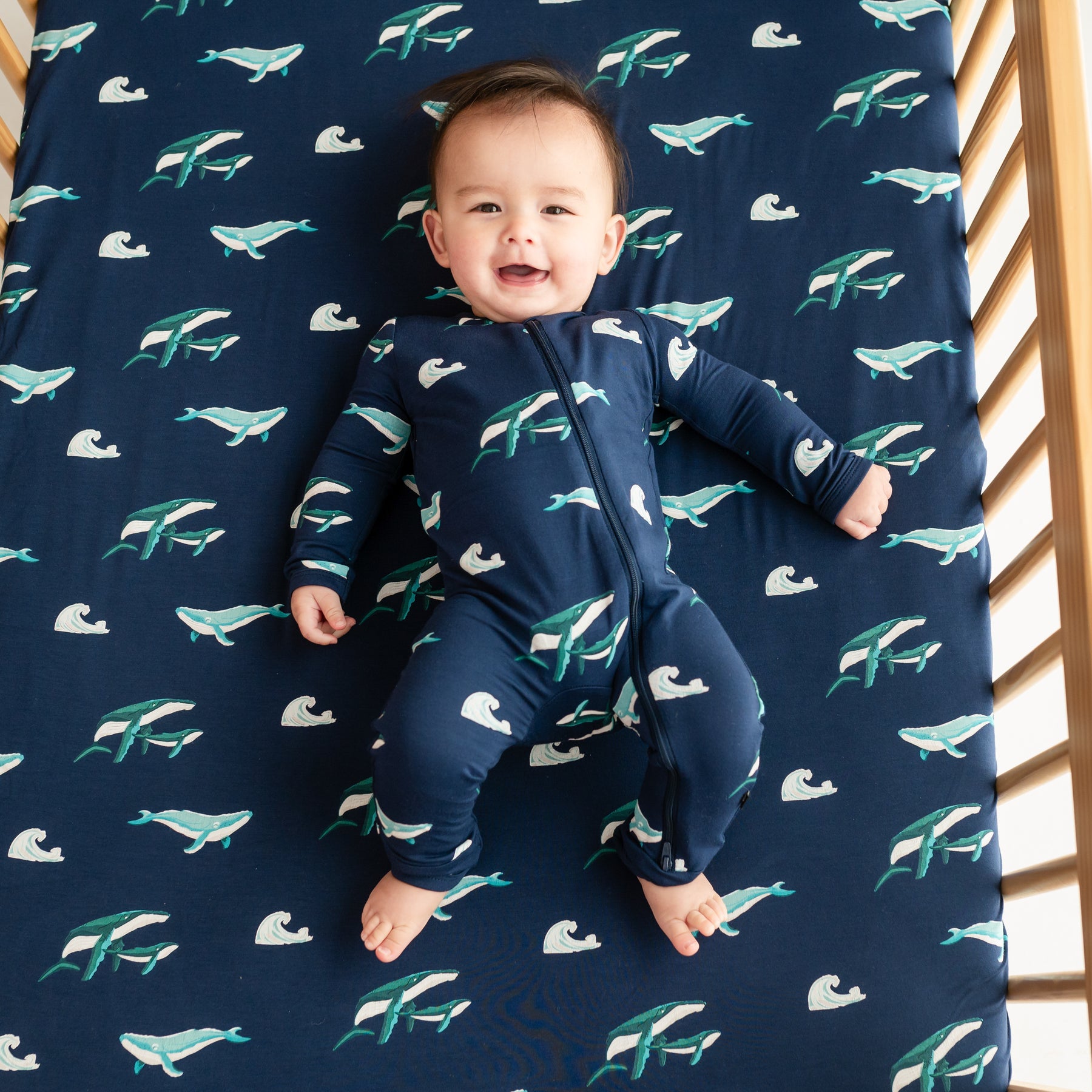 Baby wearing Zippered Romper in Humpback laying in a crib with Humpback crib sheet on