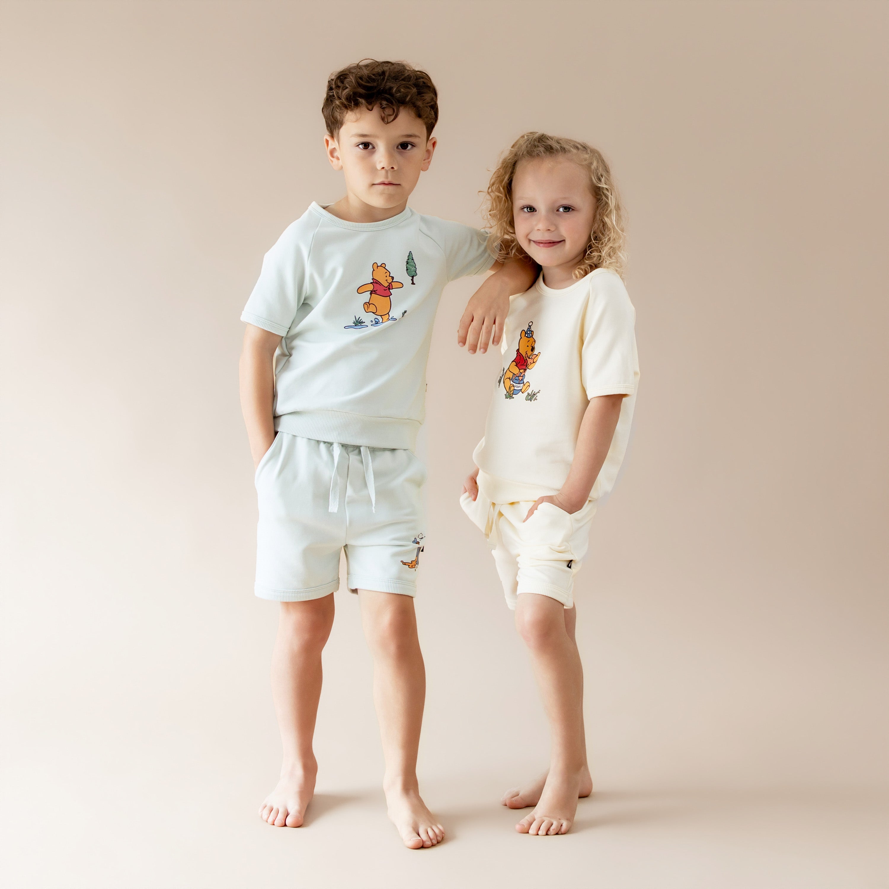 Two young children models standing side by side wearing the Short Sleeve Jogger Set in Winnie the Pooh on Dew and Winnie the Pooh in Chamomile