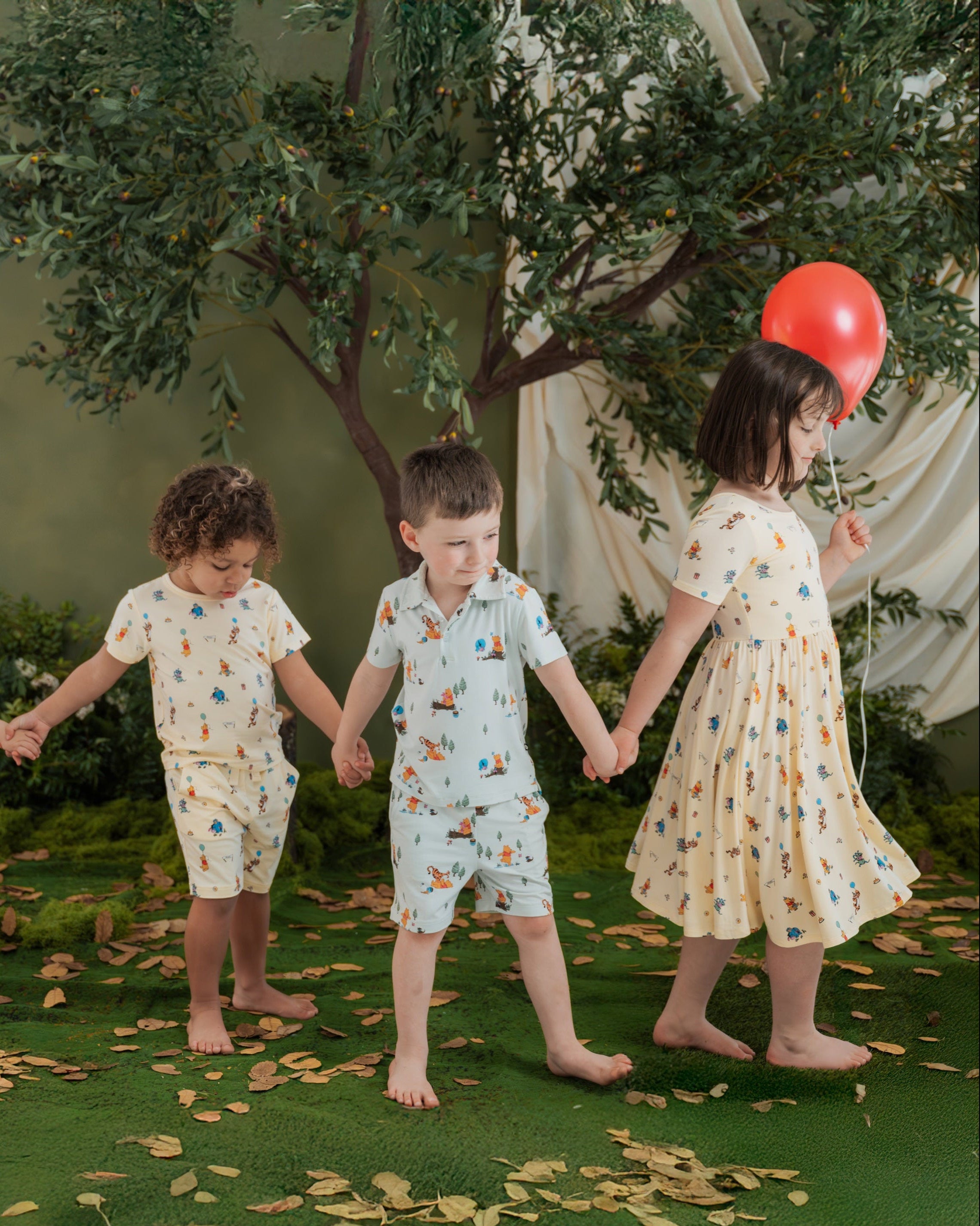 Four children holding hands in a forest setting wearing Disney Winnie the Pooh printed apparel with a red balloon.