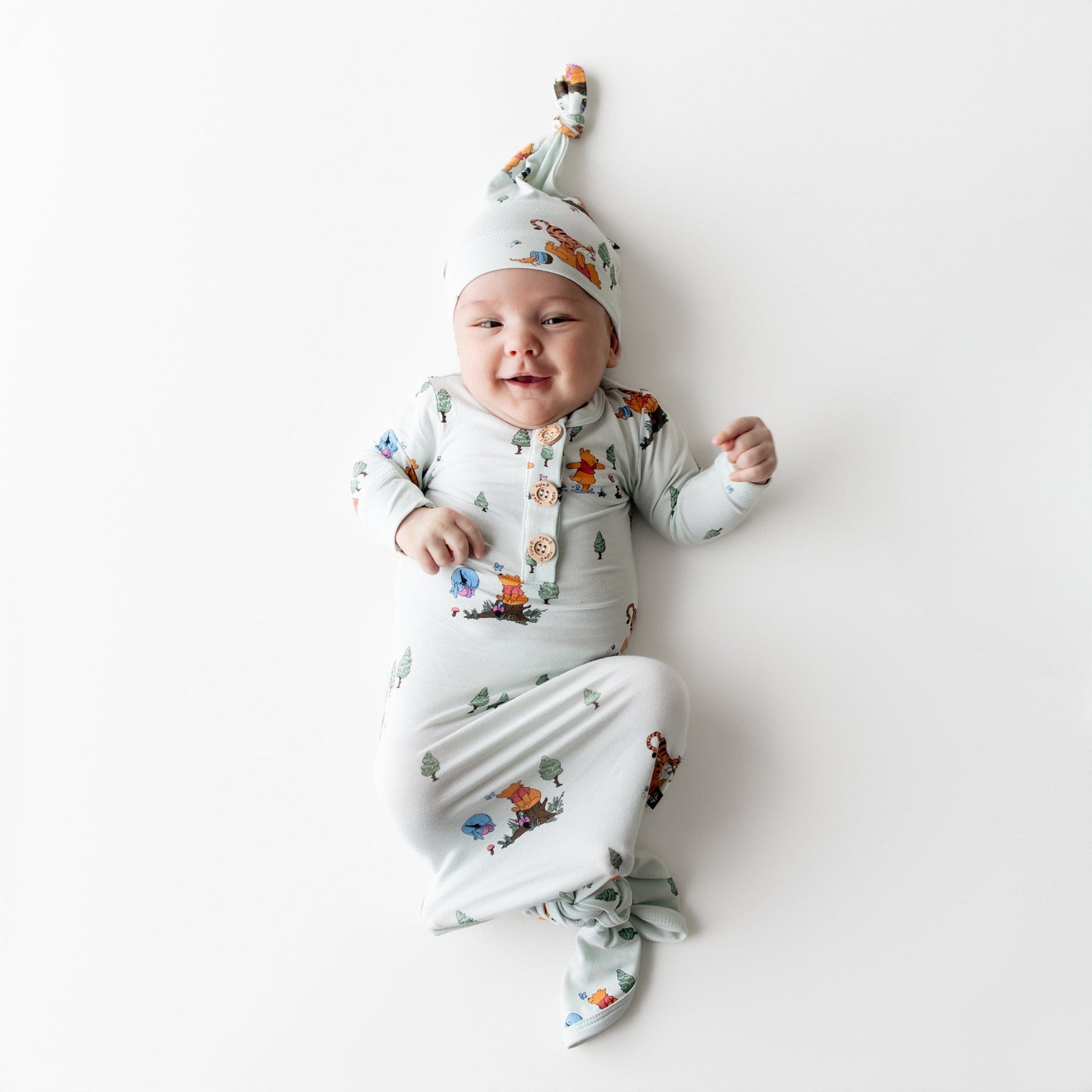 Young infant wearing the Knotted Gown with Hat Set in Winnie the Pooh Hundred Acre Wood laying on a light surface
