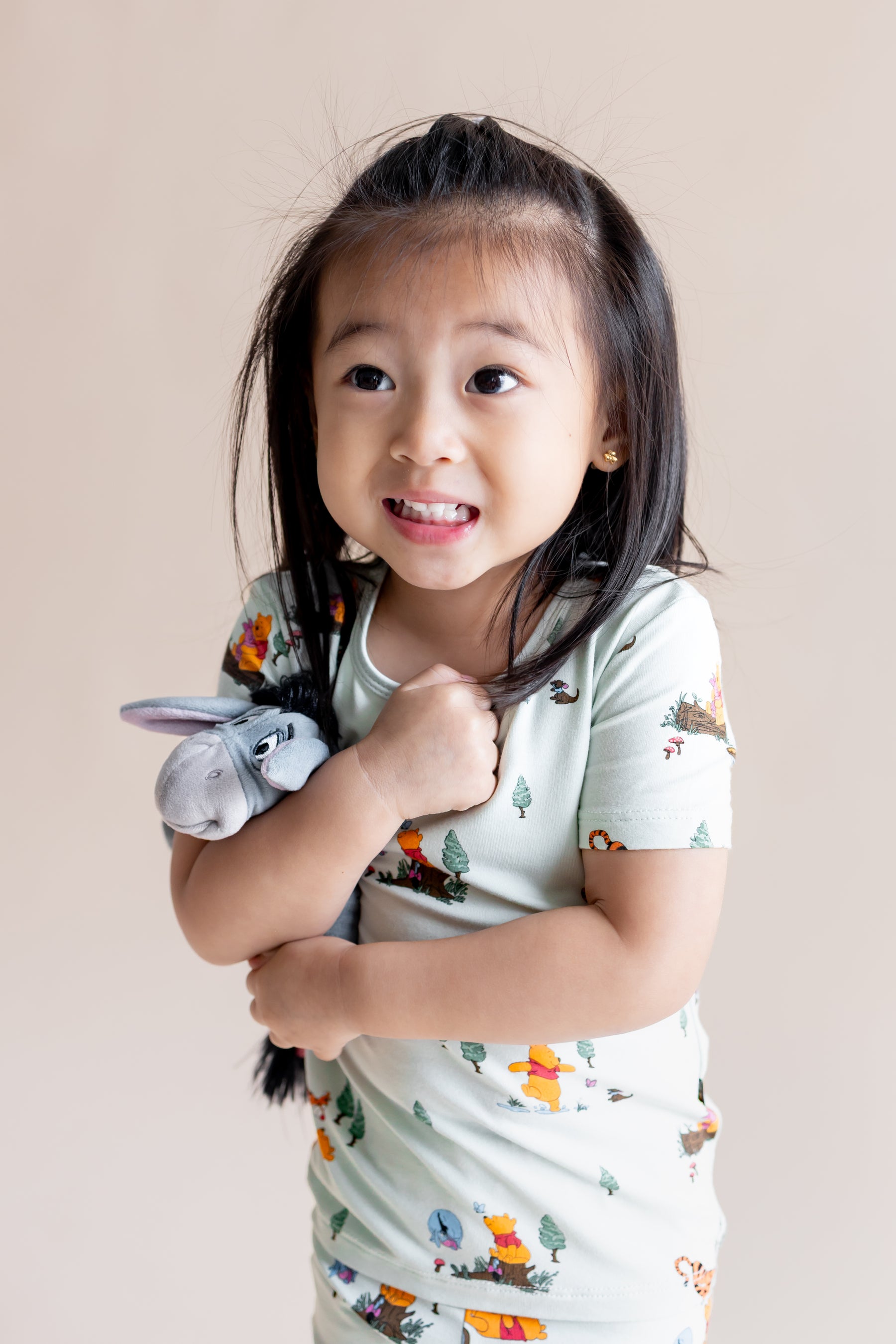 Girl holding a stuff toy wearing Short Sleeve Pajamas in Winnie the Pooh Hundred Acre Wood print on a cream background