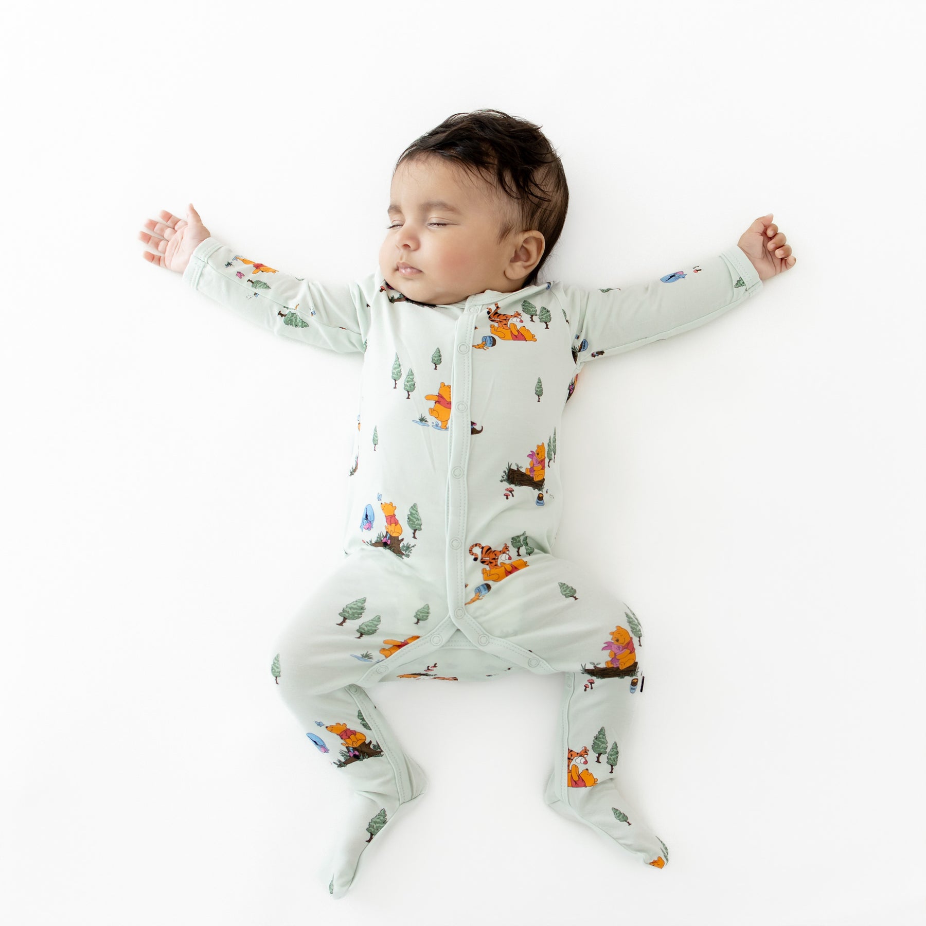 Sleeping infant stretched out wearing the Footie in Winnie the Pooh Hundred Acre Wood on a white surface