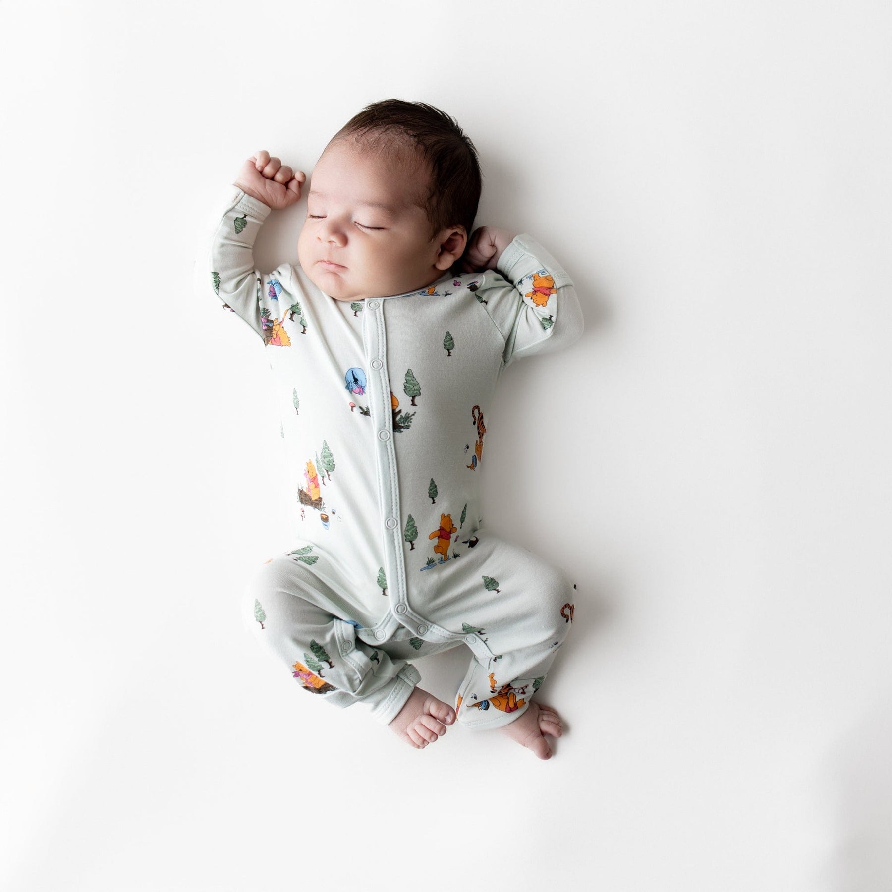 Sleeping newborn laying on a light neutral surface wearing the Romper in Winnie the Pooh Hundred Acre Wood