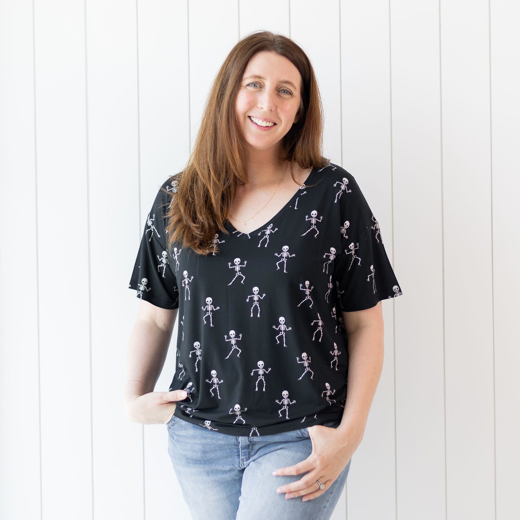 Smiling female wearing the Women's Relaxed Fit V-Neck in Skeleton paired with light wash jeans standing in front of a white paneled wall