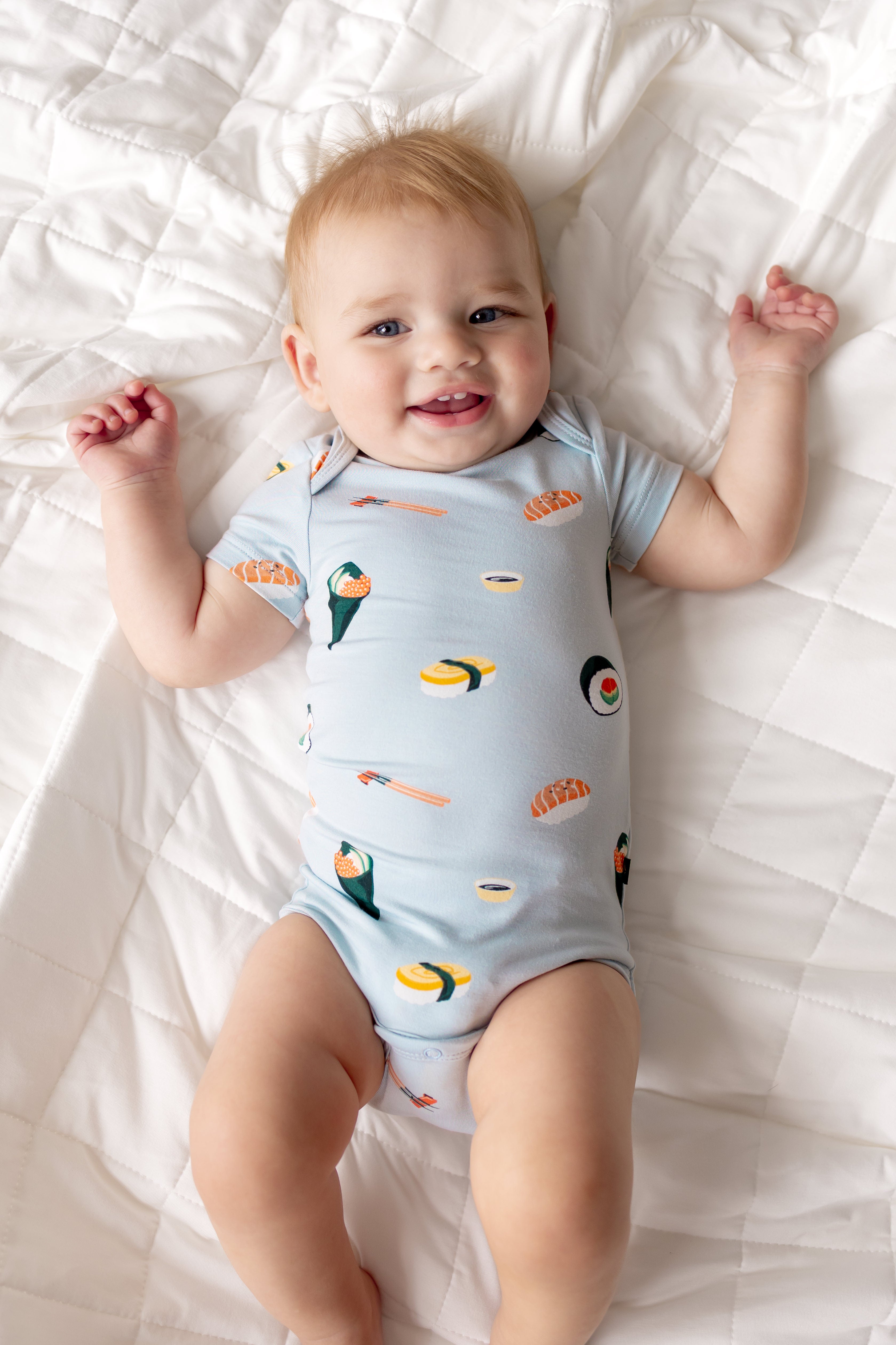 Baby lying on white sheet wearing Blue Bodysuit in Sushi print
