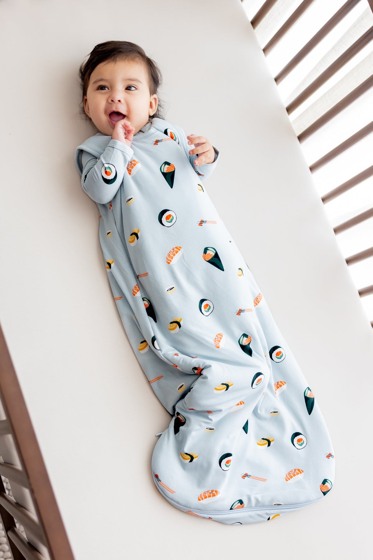 Baby laying in crib on white sheets with his hand to his mouth in ice sushi sleep bag