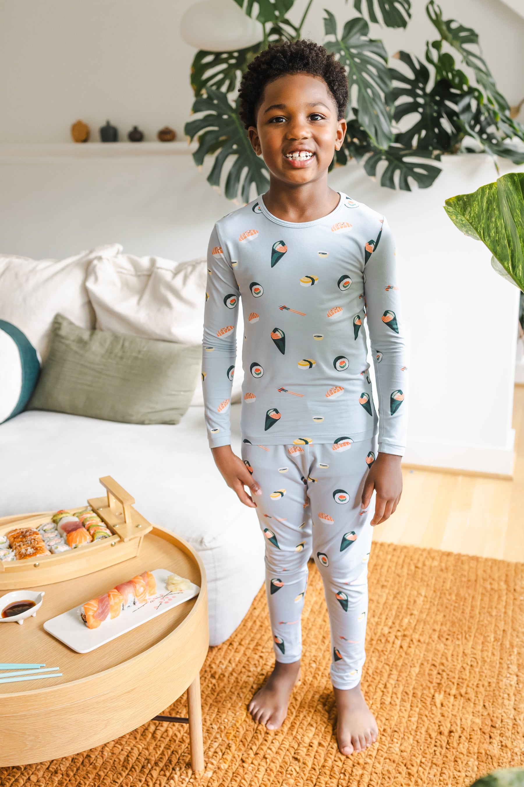 Child smiling in ice sushi pajama se, standing next to a table of sushi with a plant in the background