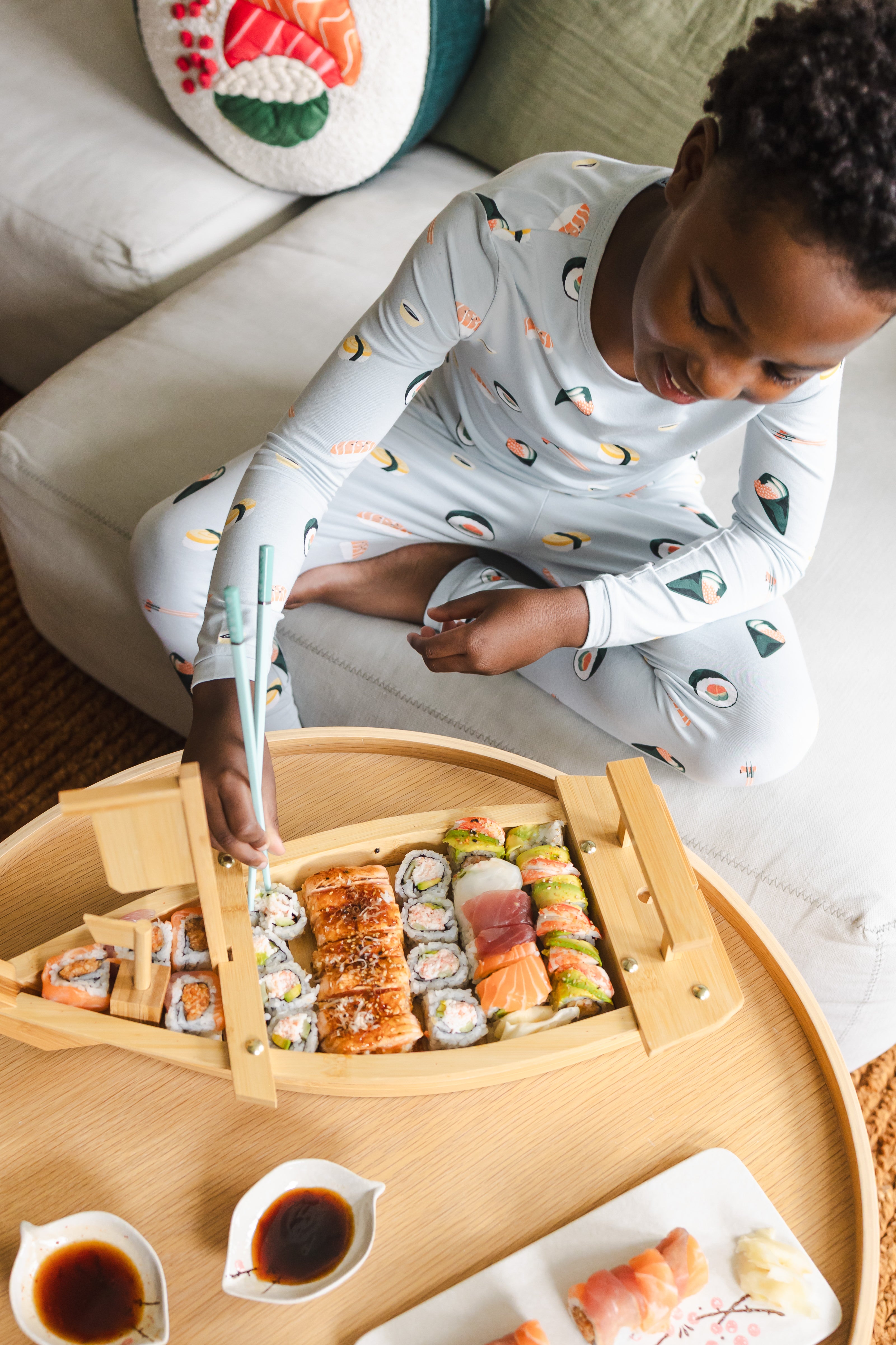 Child sitting on the couch looking down at the sushi he is eating with chop sticks