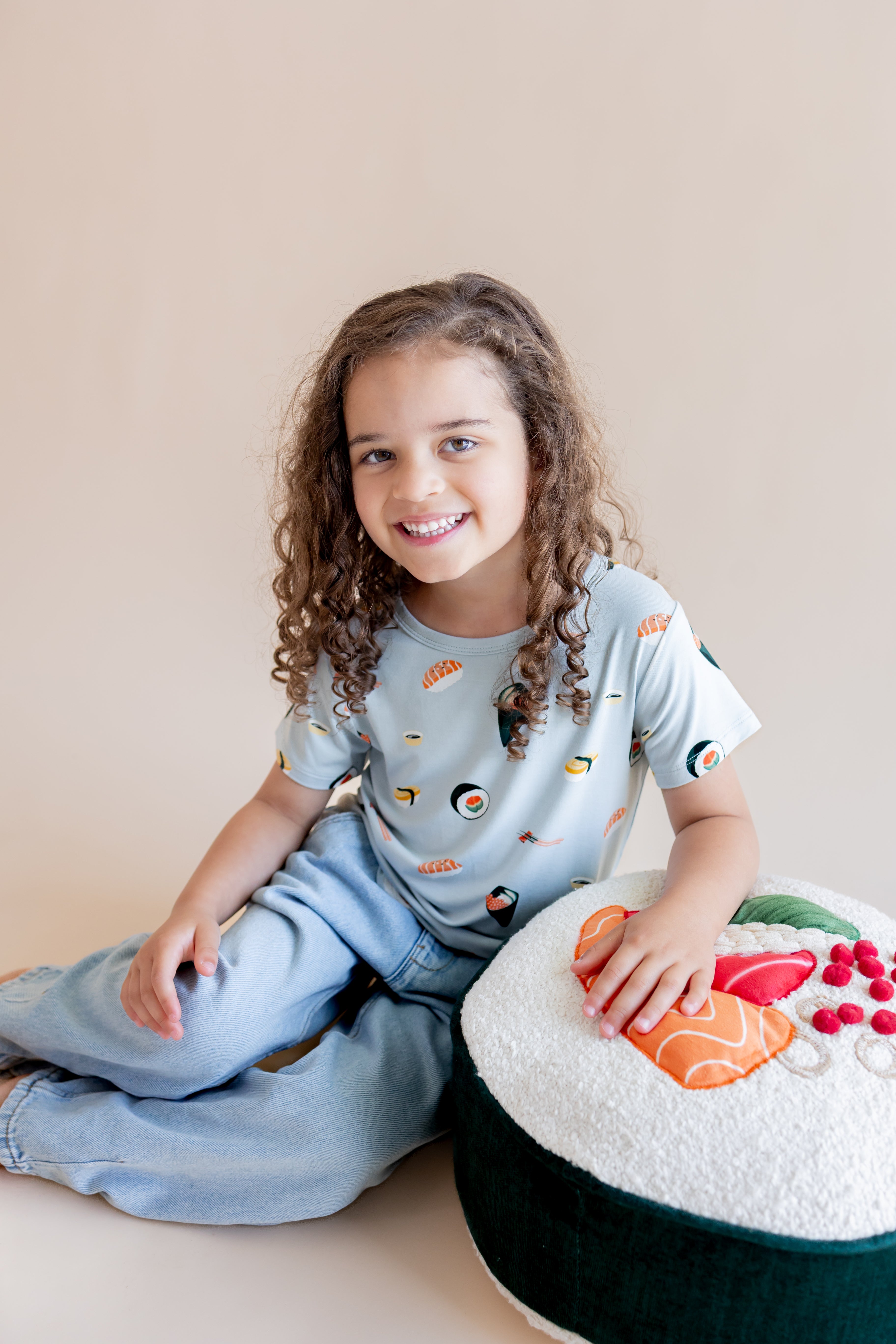 Child smiling wearing ice sushi tee in pants, sitting cross-legged with a plate of play sushi beside them