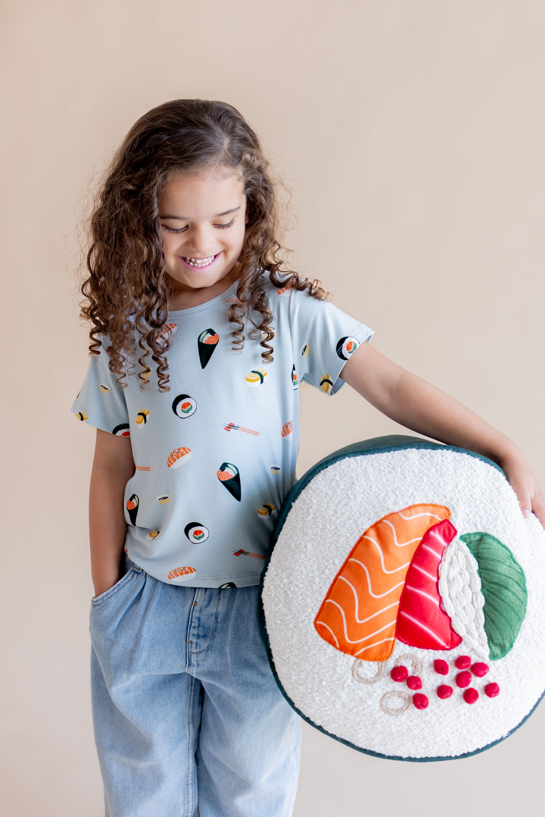 Child wearing ice sushi tee while looking down at a play sushi pillow she is holding on her hip