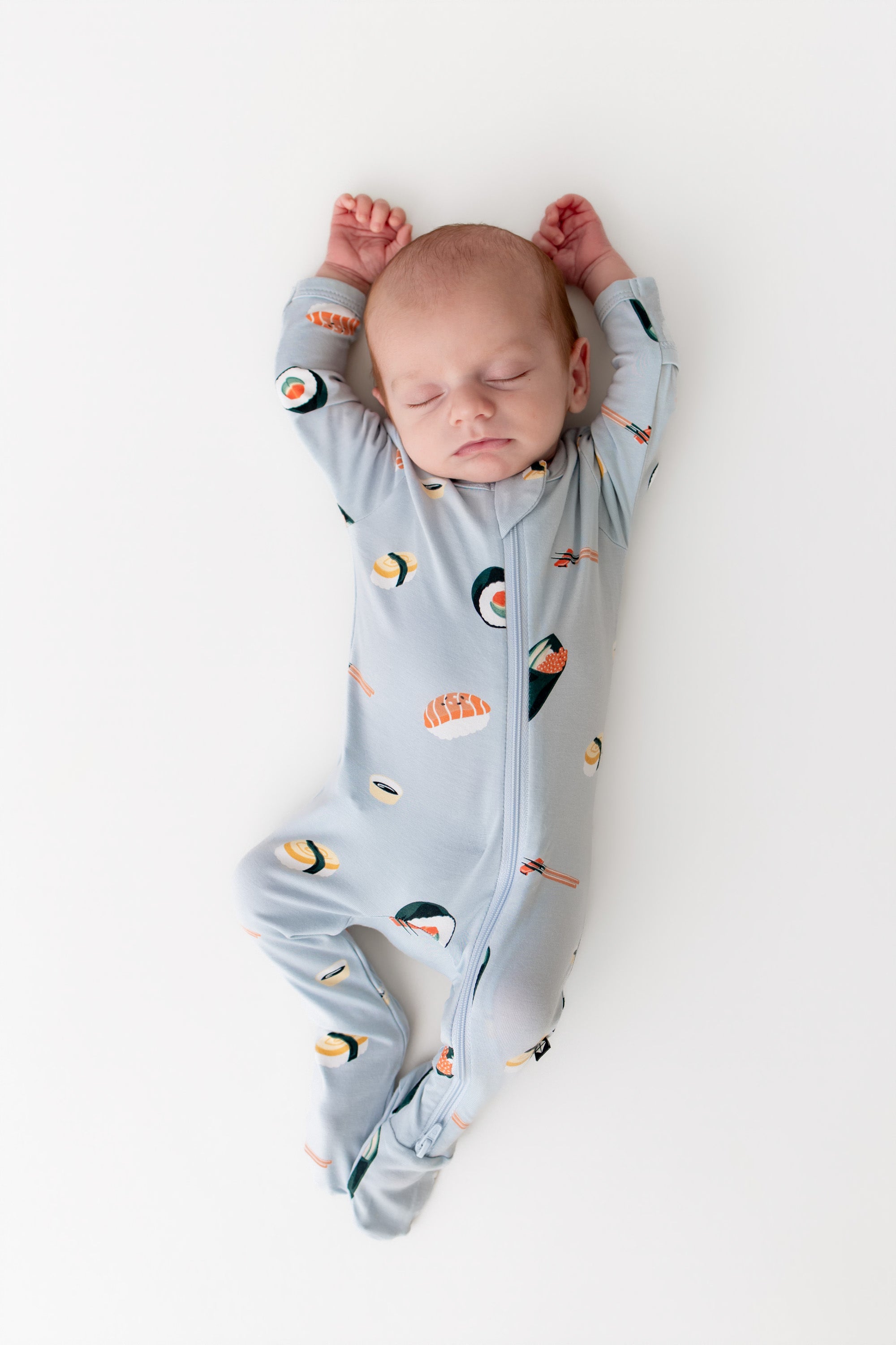 Baby sleeping with their arms up in ice sushi zipper footie on a white background