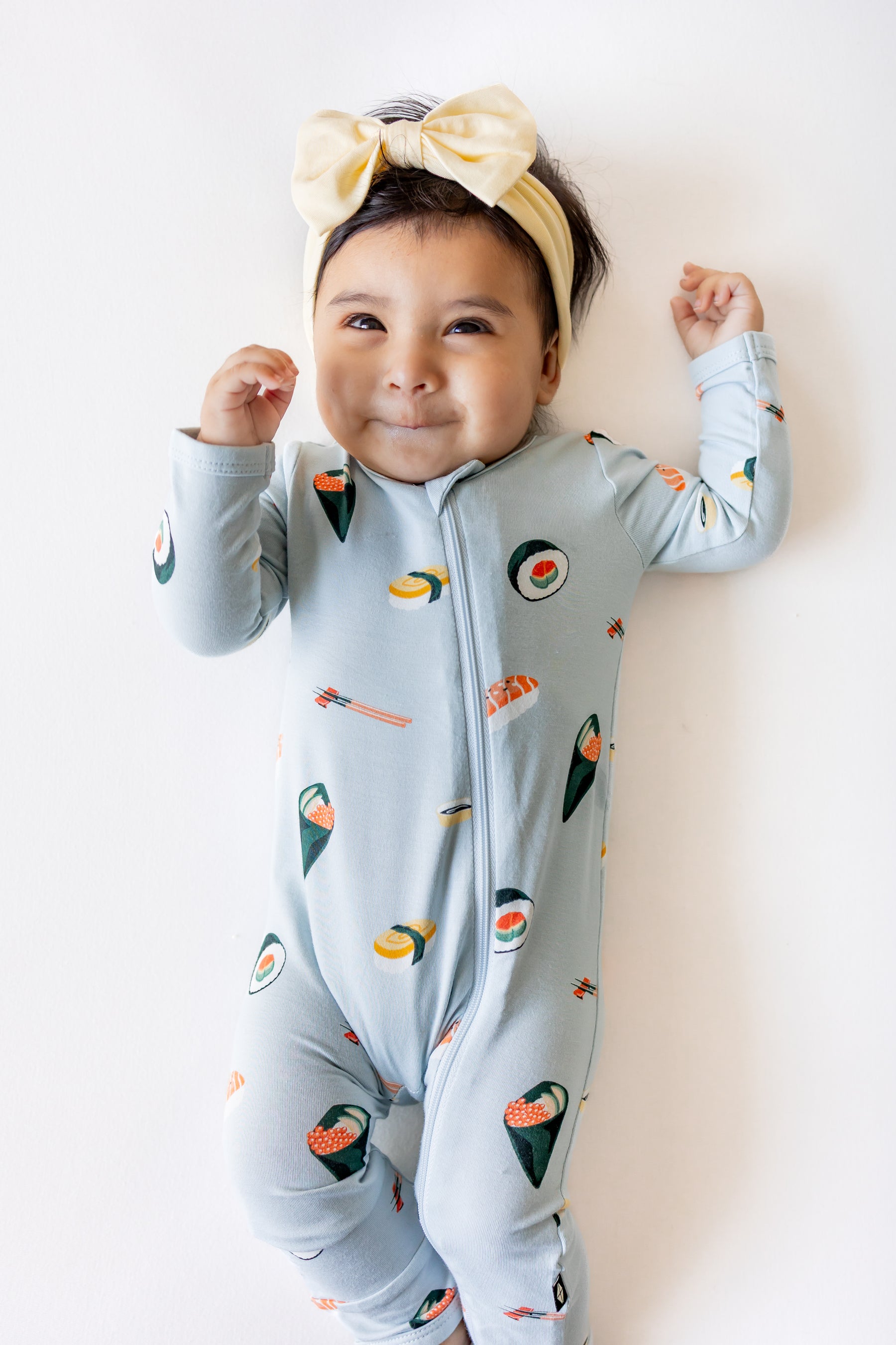 Baby laying down with a white background wearing ice sushi pajama set and  a yellow bow