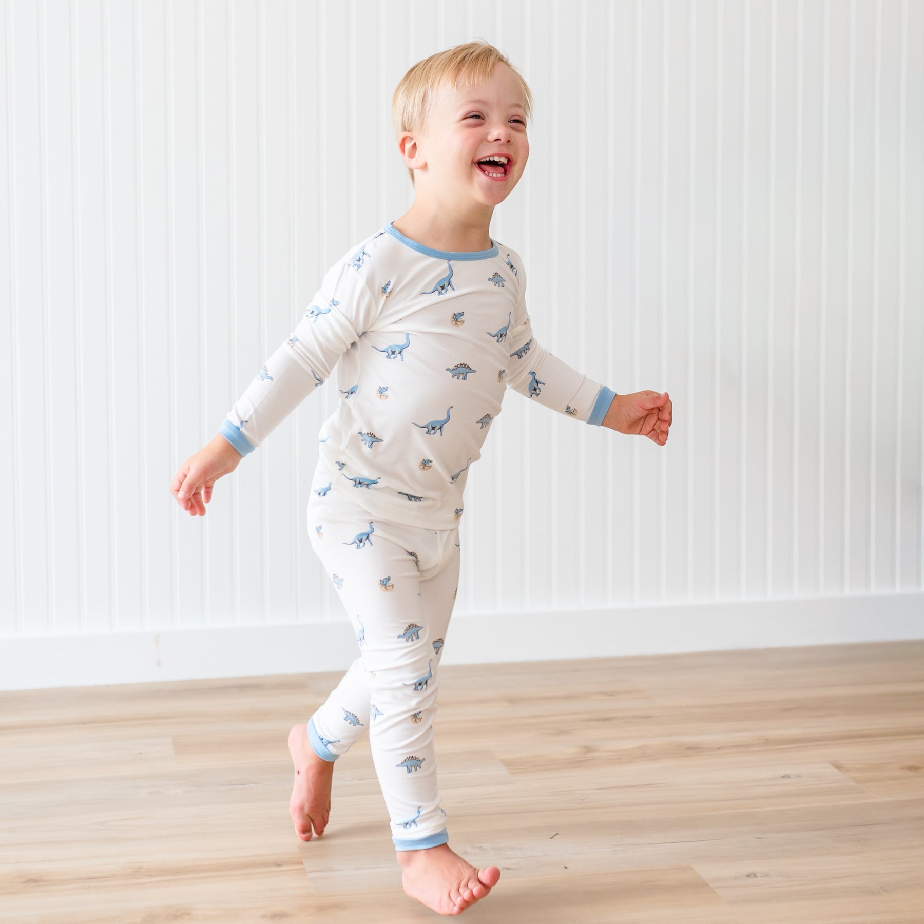Child wearing a long-sleeve pajama set with a dinosaur pattern on a wooden floor.