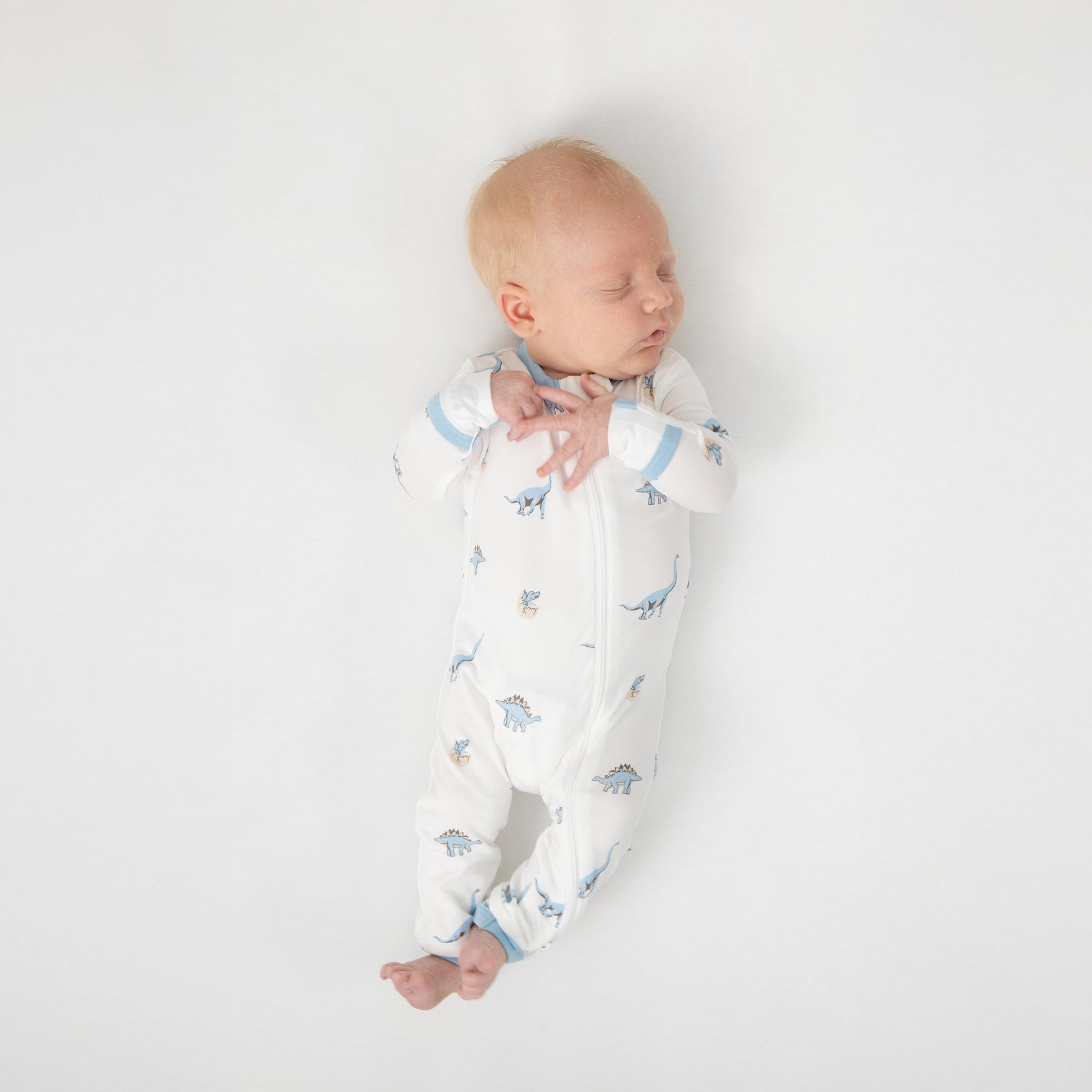 Sleeping newborn laying on a white crib sheet wearing the Zippered Romper in Jurassic