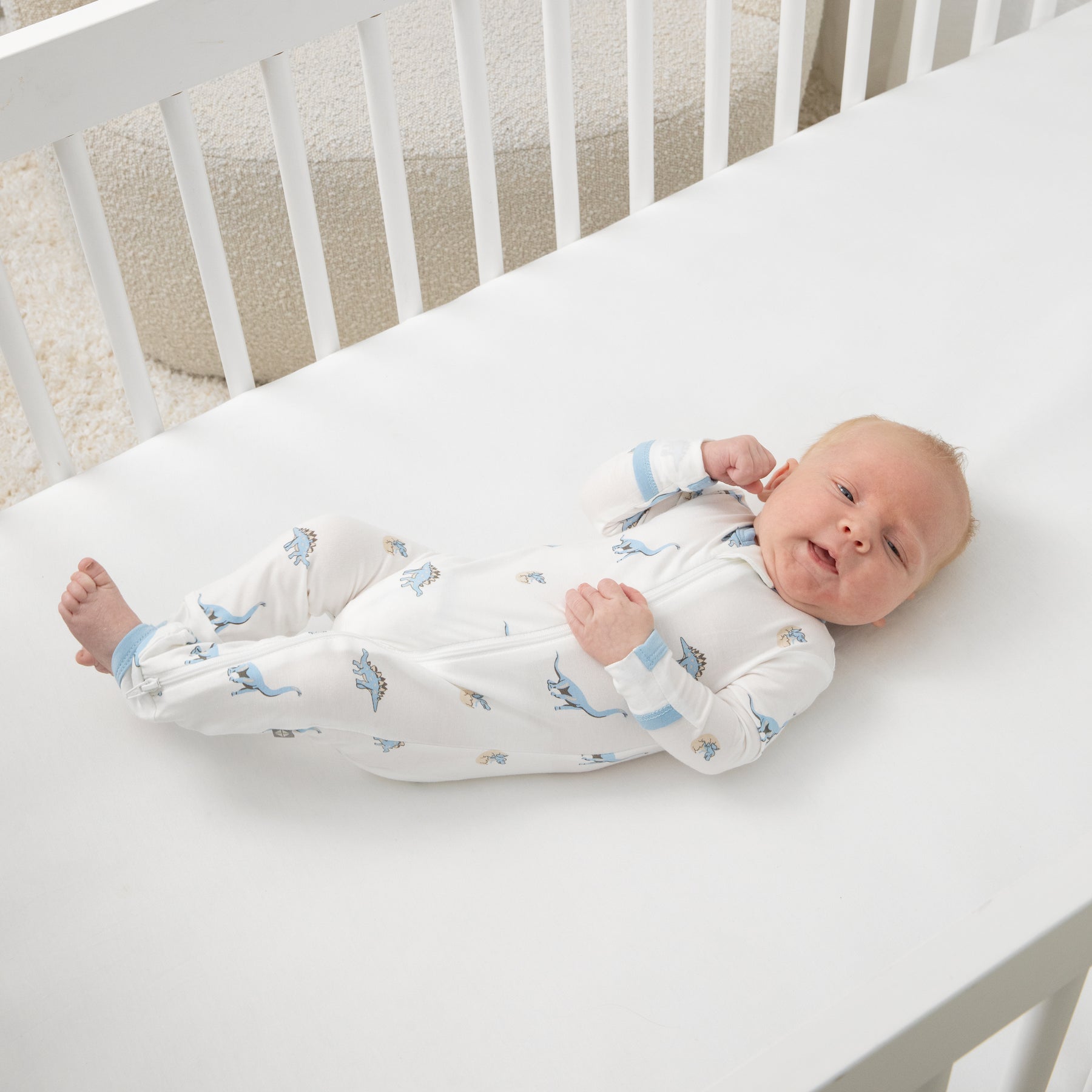 Newborn baby laying in a crib on a white crib sheet wearing the Zippered Romper in Jurassic
