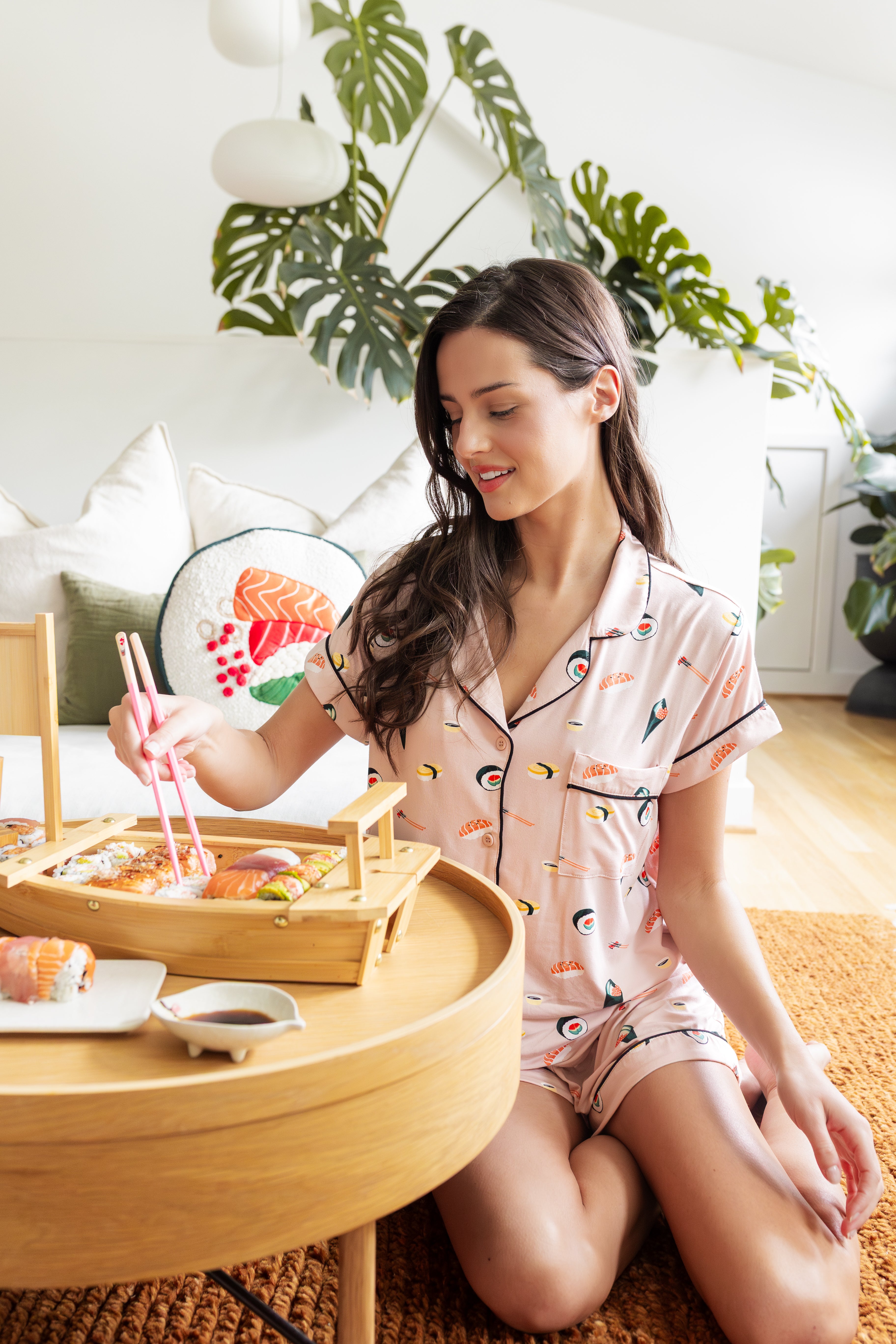 Woman in pink sushi themed pajamas sitting on the floor with a tray of food in a cozy room.