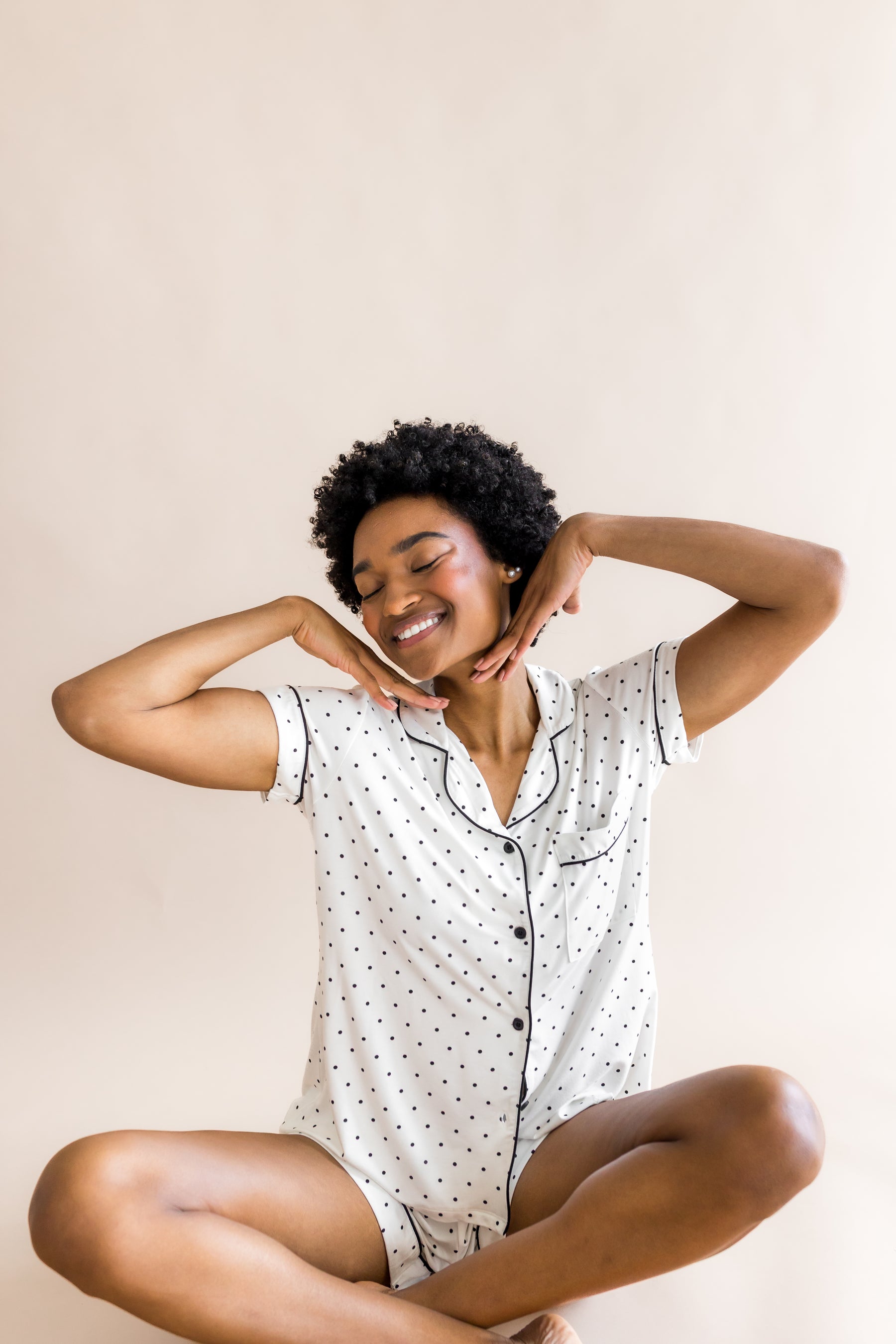 Woman sitting wearing a polka dot short sleeve pajama set with black piping on a beige background
