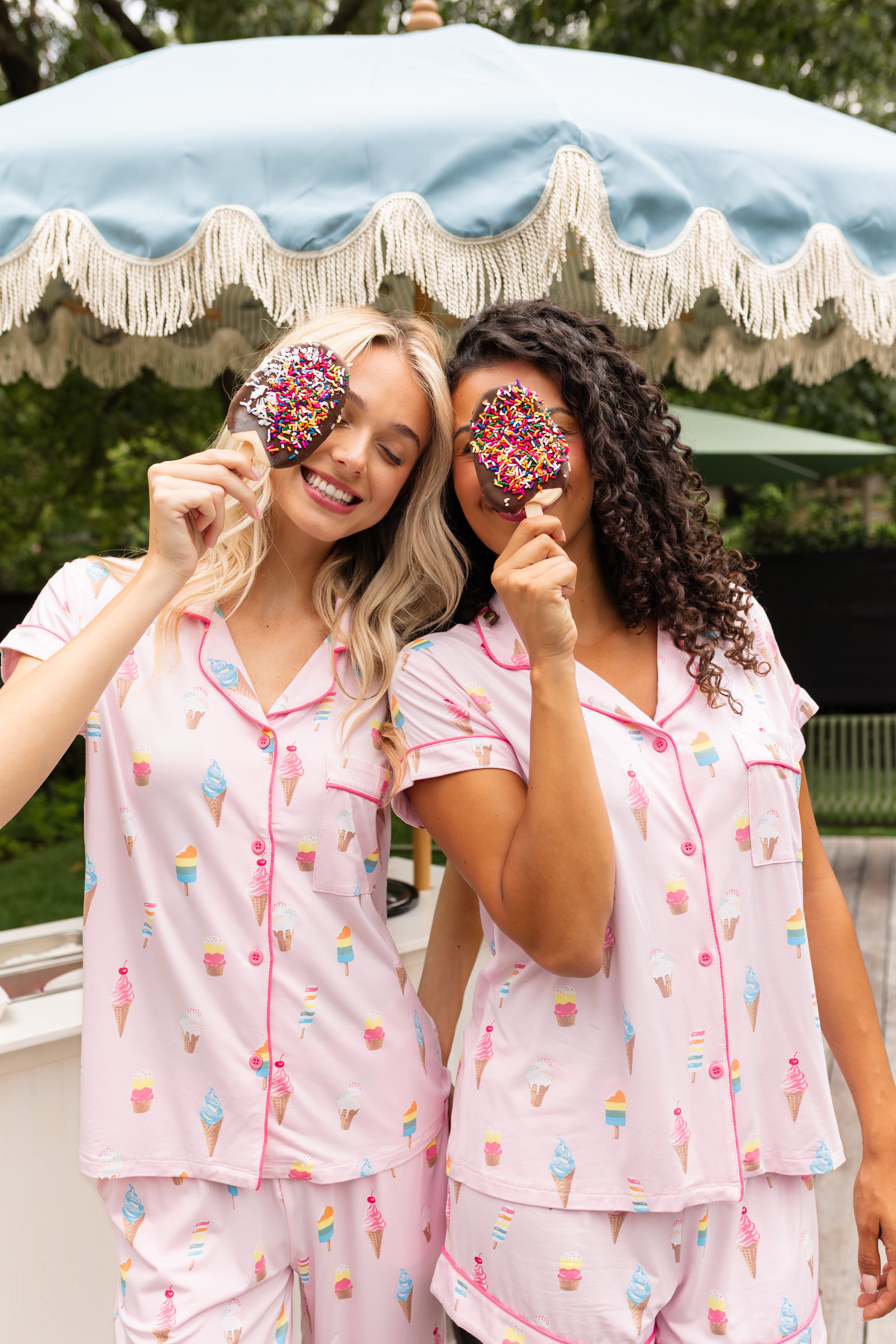 Two Models smiling in Women's Short Sleeve Pajama Set and Women's Short Sleeve Cropped Pajama Set in Sakura Ice Cream print outdoors with an ice cream stick in front of an umbrealla