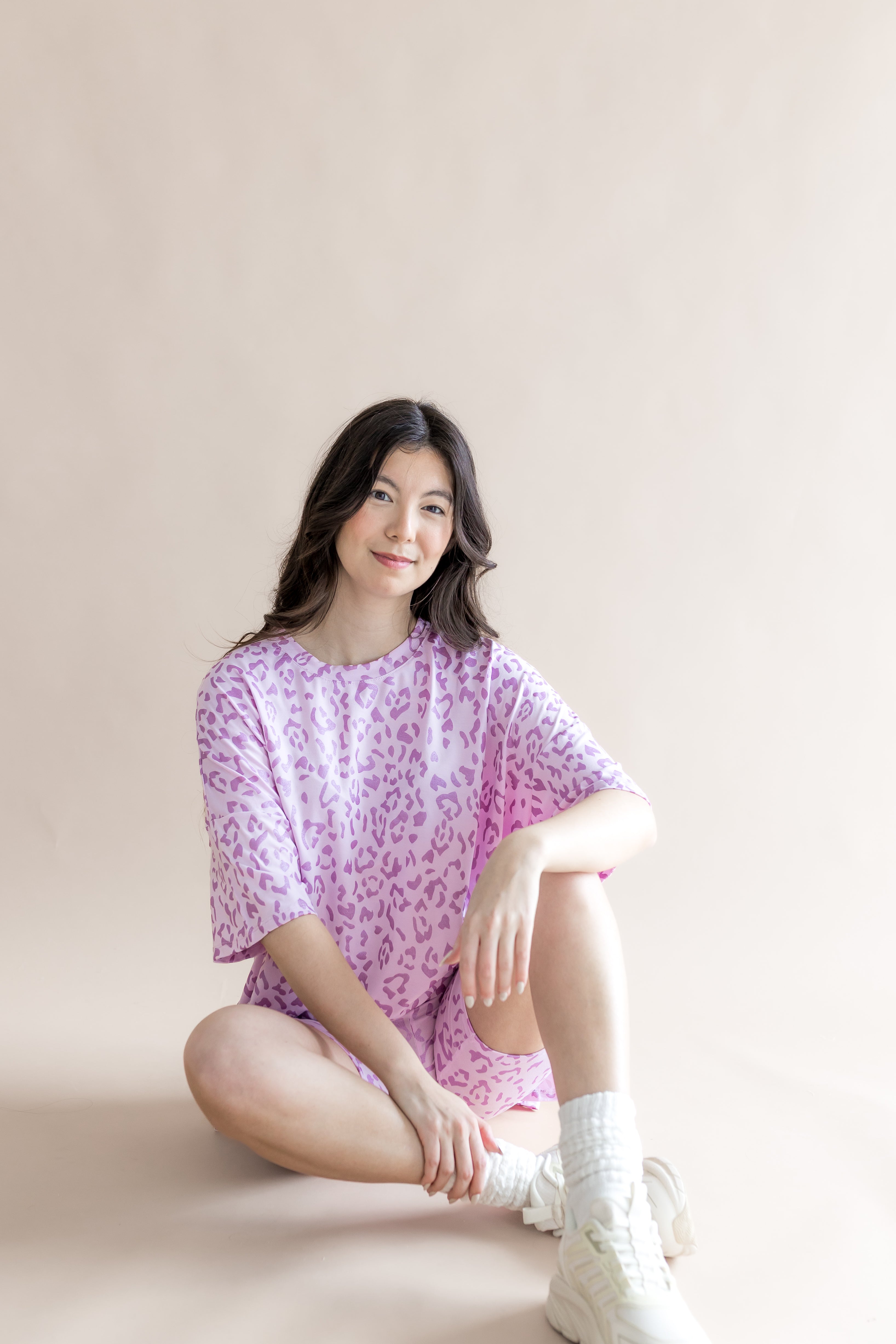 Model sitting in Women’s Biker Short Set in Thistle Leopard print with white socks and shoes on a beige background