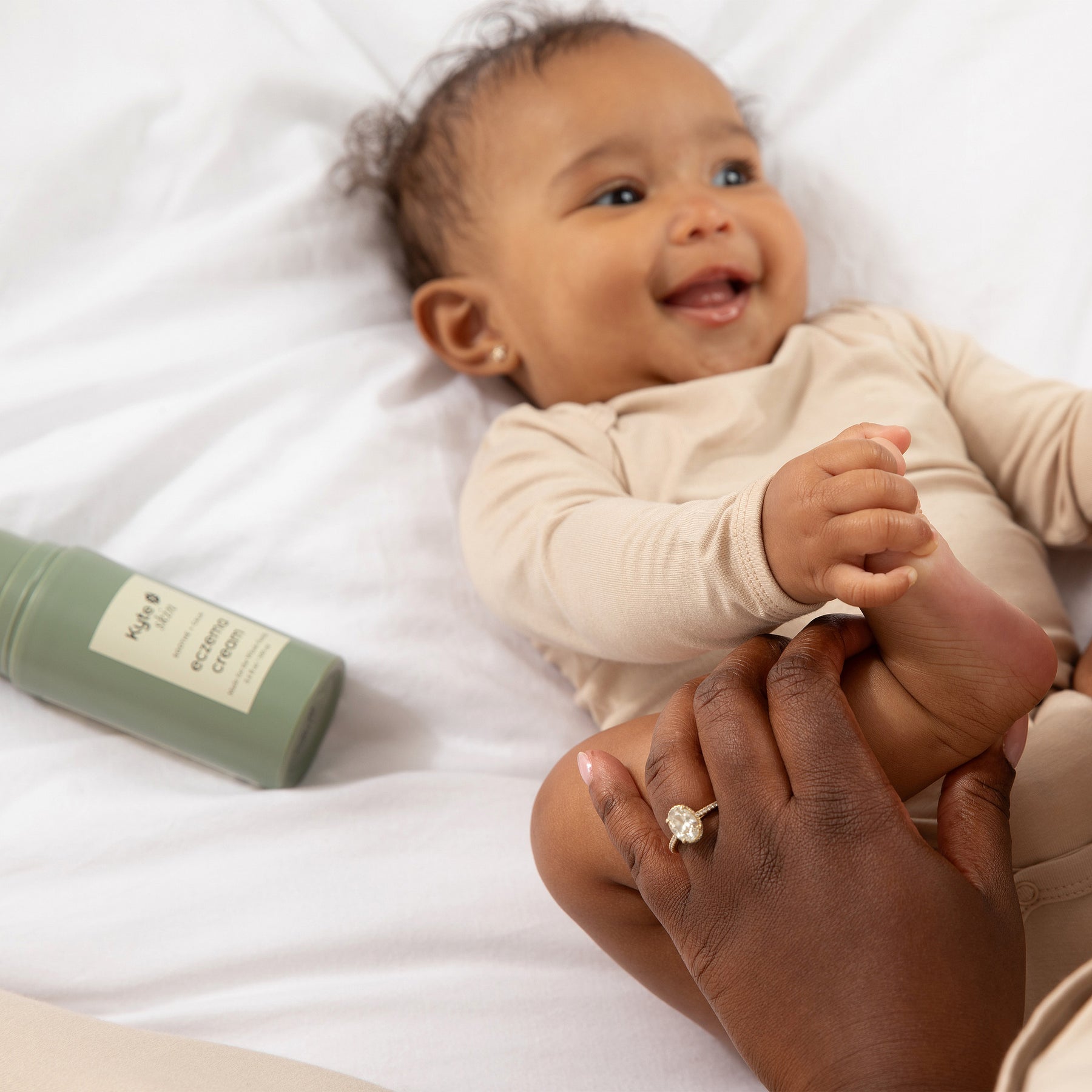 Baby lying on a white surface with a bottle of lotion next to it, held by an adult hand.