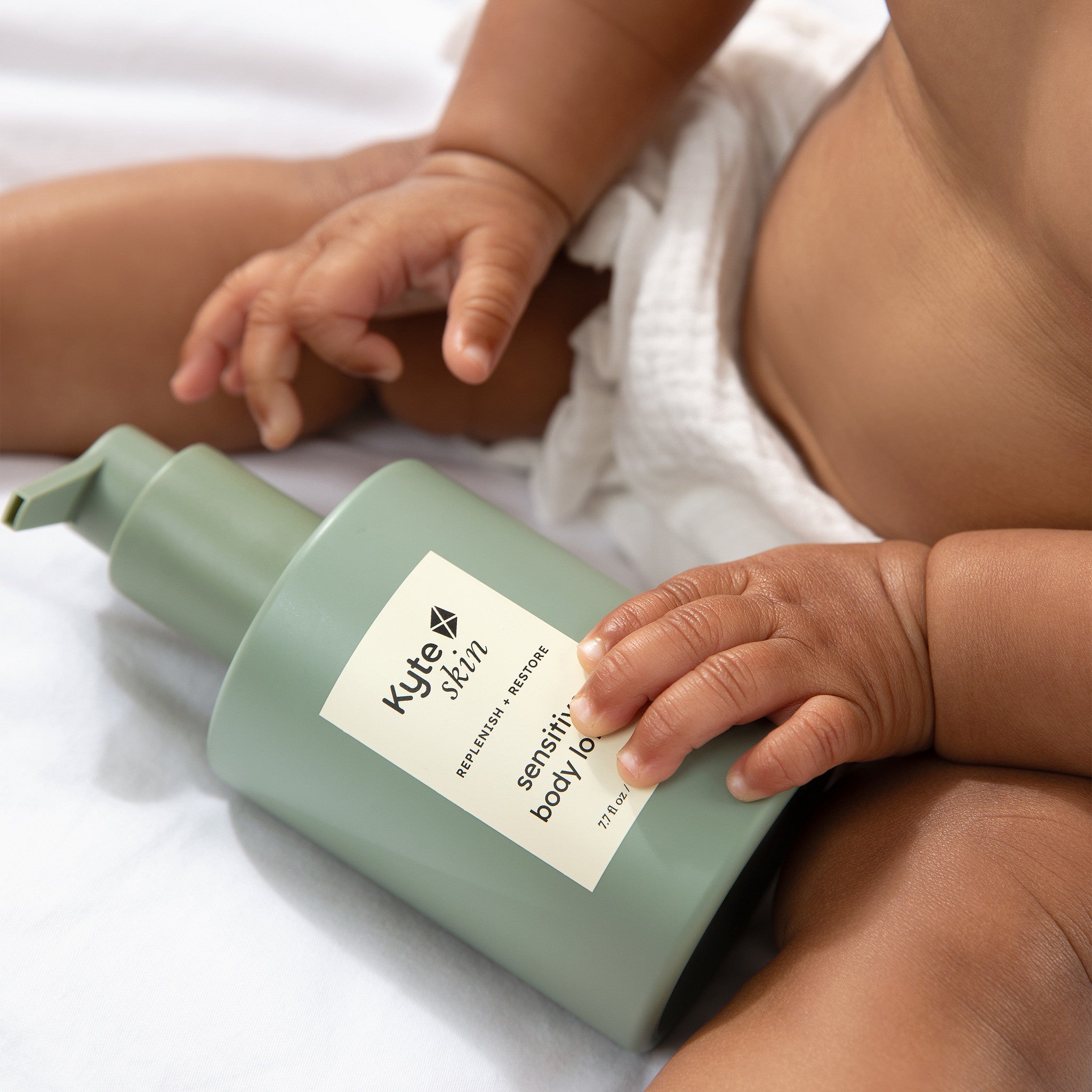Baby's hand touching a bottle of Kyte Skin sensitive body balm on a white surface
