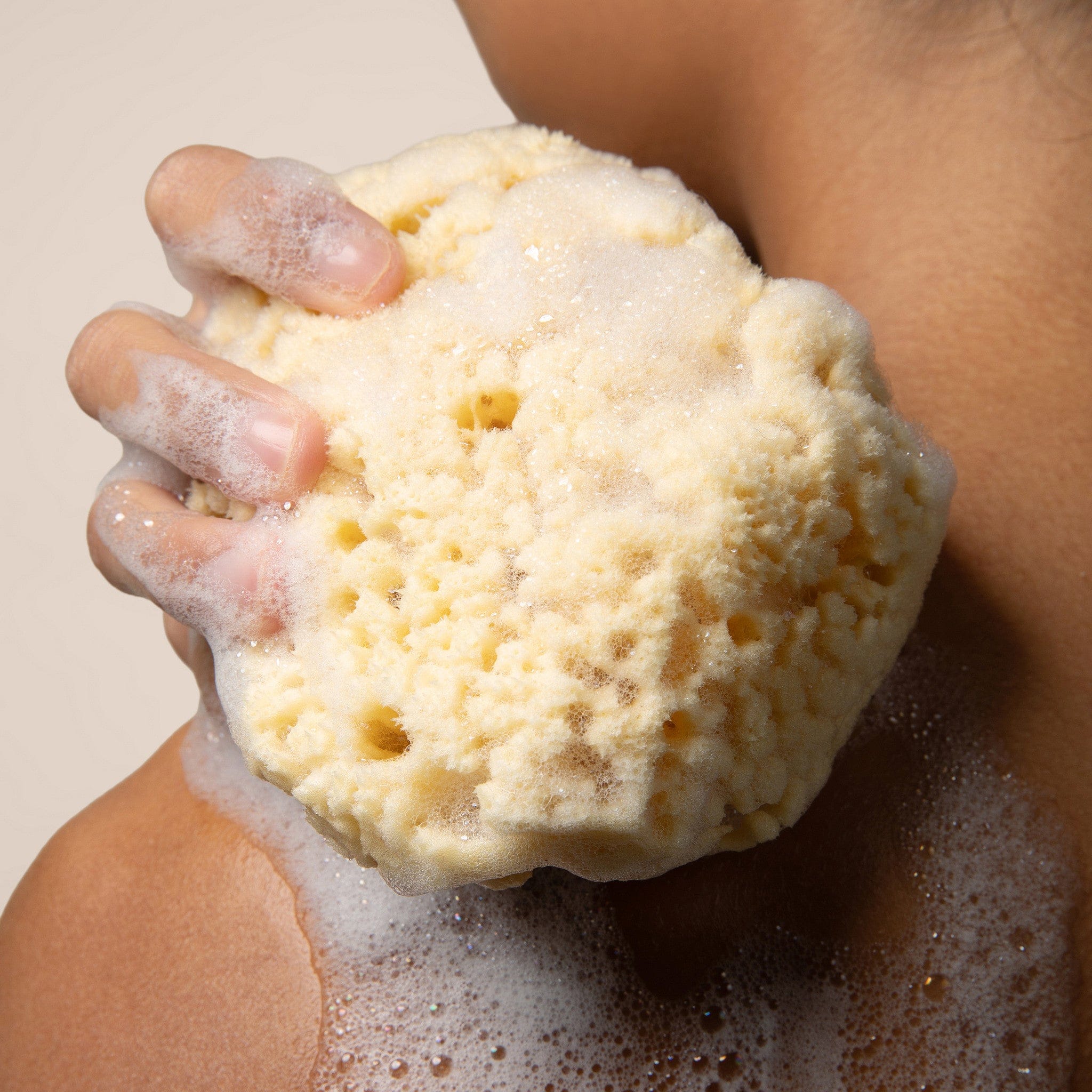 Hand holding a sudsy yellow bath sponge against a neutral background