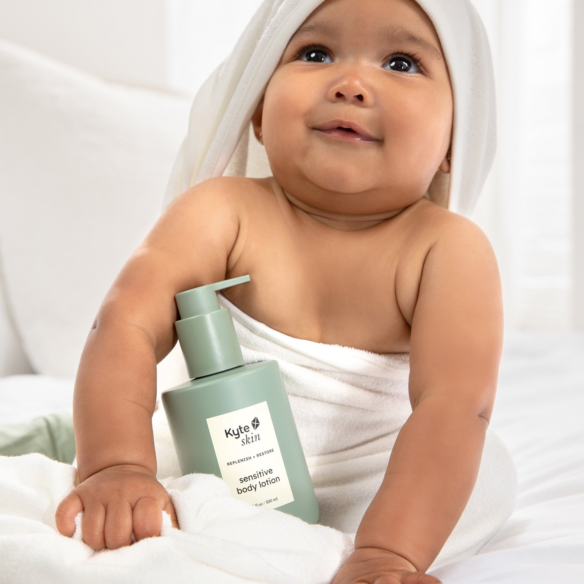 Baby holding a bottle of Kyte Skin sensitive body lotion with a white towel on head.