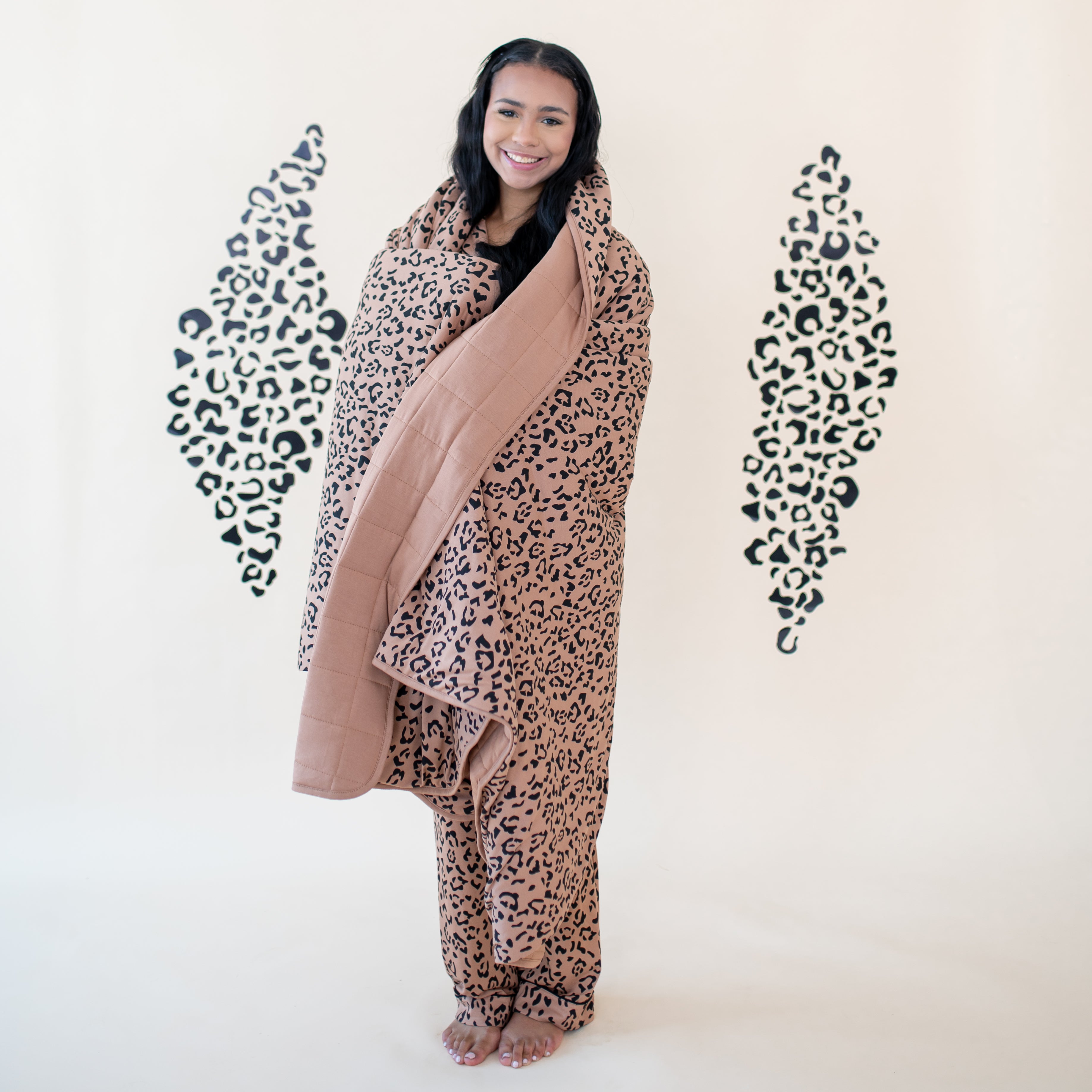 Smiling female model with the Adult Quilted Blanket in Latte Leopard 2.5 wrapped around her in front of a leopard print backdrop