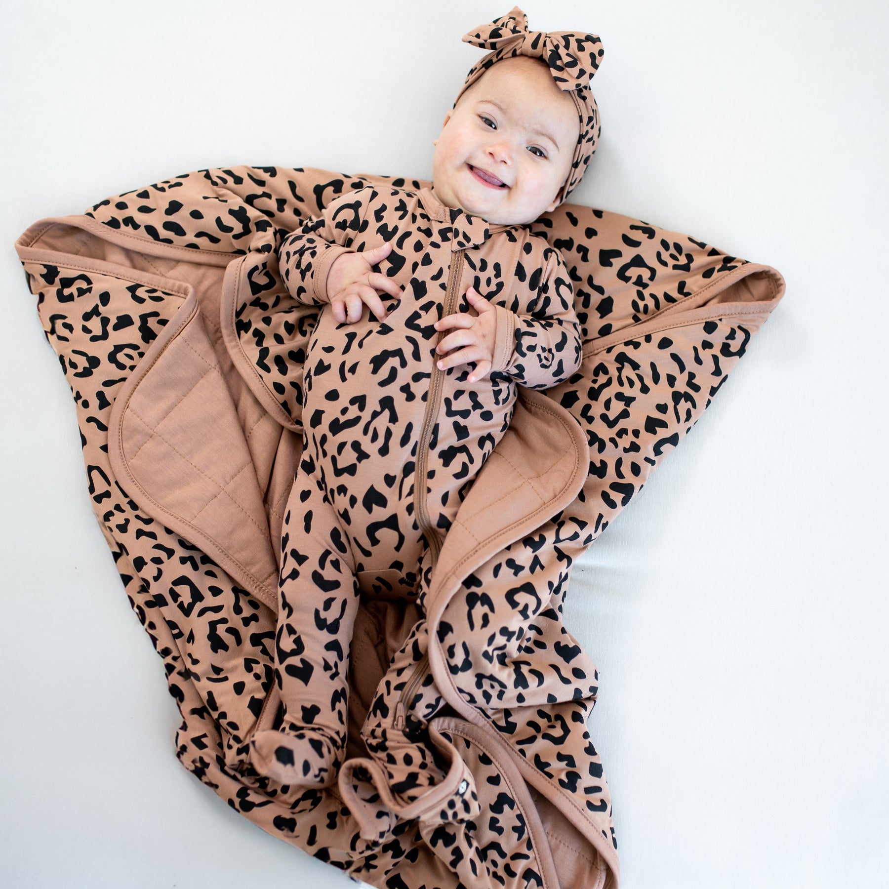 Infant laying on Baby Blanket in Latte Leopard wearing matching zippered footie pajamas and bow headband
