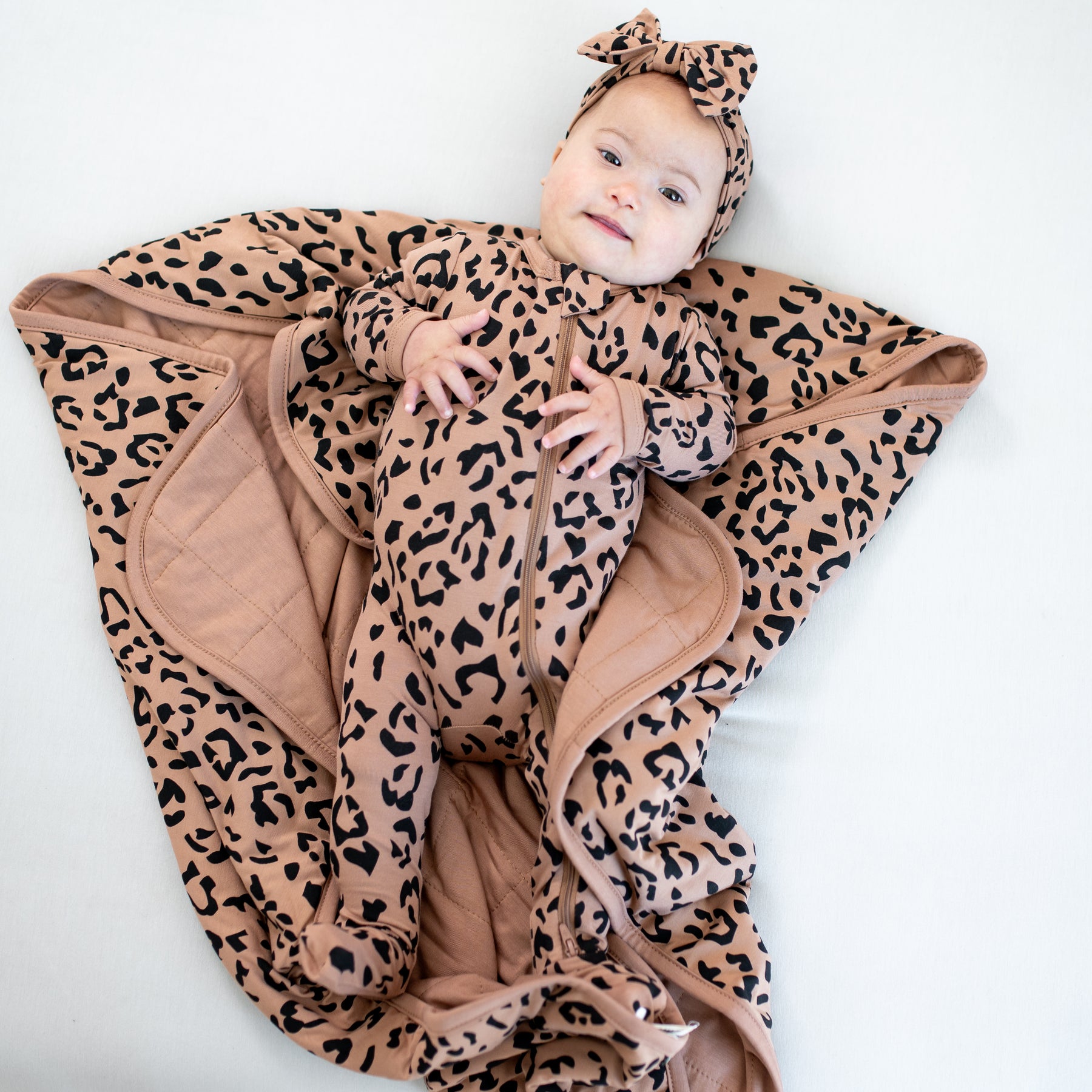 Infant wearing the Bow Headband in Latte Leopard paired with a matching zippered footie laying on a Latte Leopard baby blanket