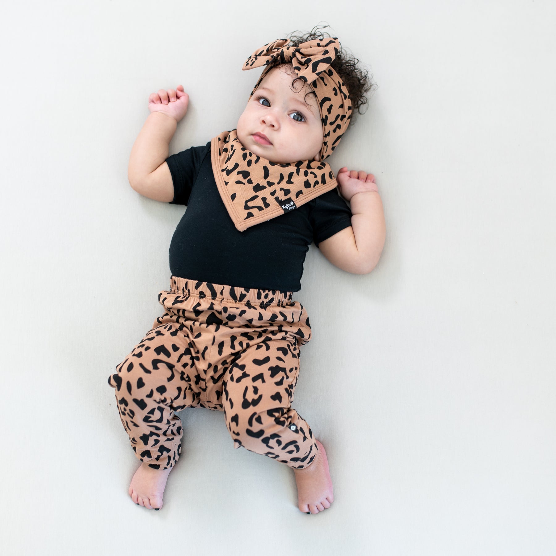 Infant laying on a light colored blanket wearing the Bib in Latte Leopard paired with matching bow headband, pants and a Midnight bodysuit