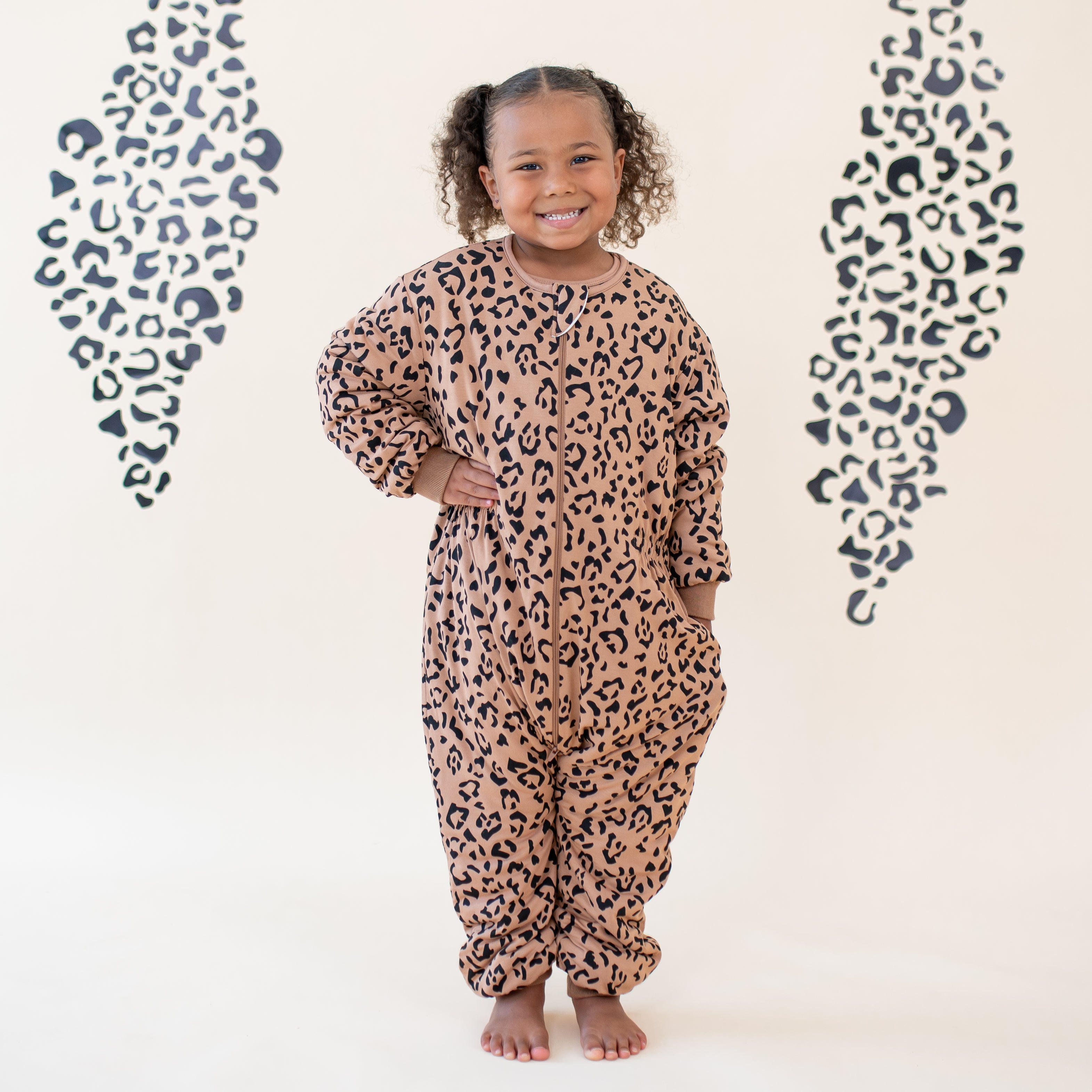 Smiling young girl wearing the Cozy Playsuit in Latte Leopard with one hand in the pocket and the other on her hip