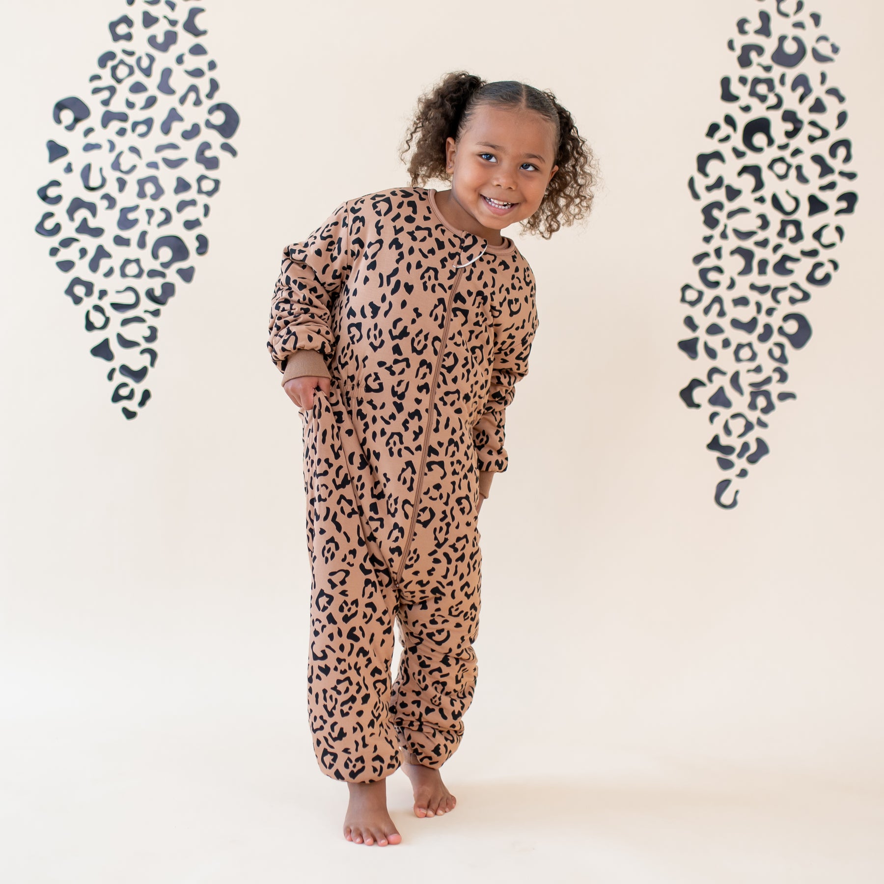 Young girl walking in the Cozy Playsuit in Latte Leopard in front of a neutral leopard backdrop