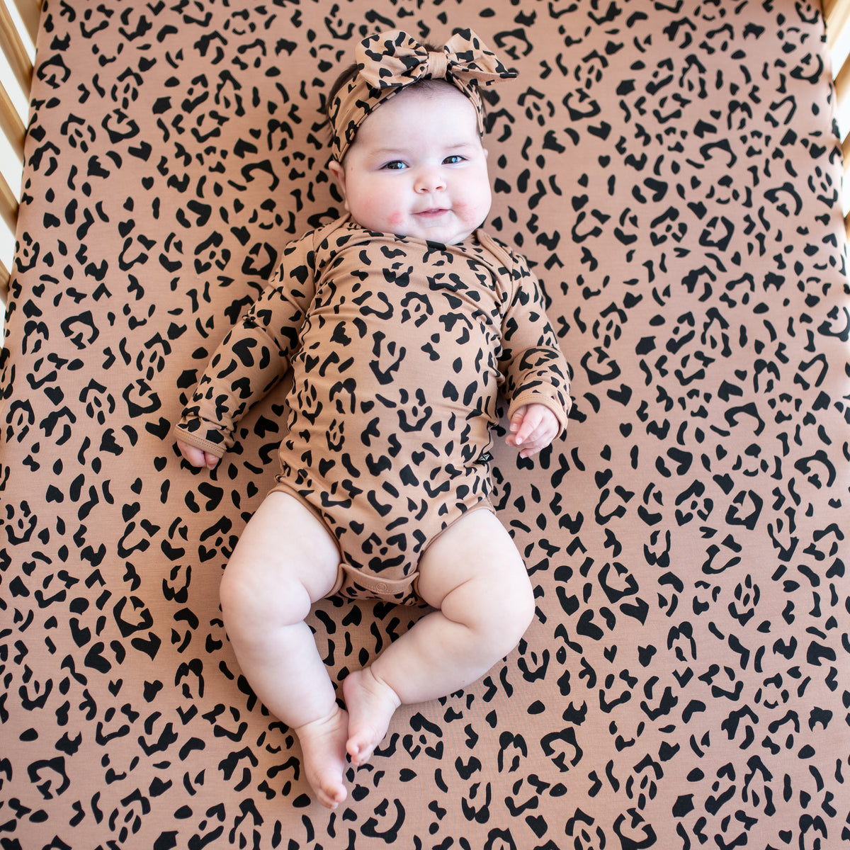 Infant laying on a Crib Sheet in Latte Leopard wearing the matching long sleeve bodysuit and bow headband