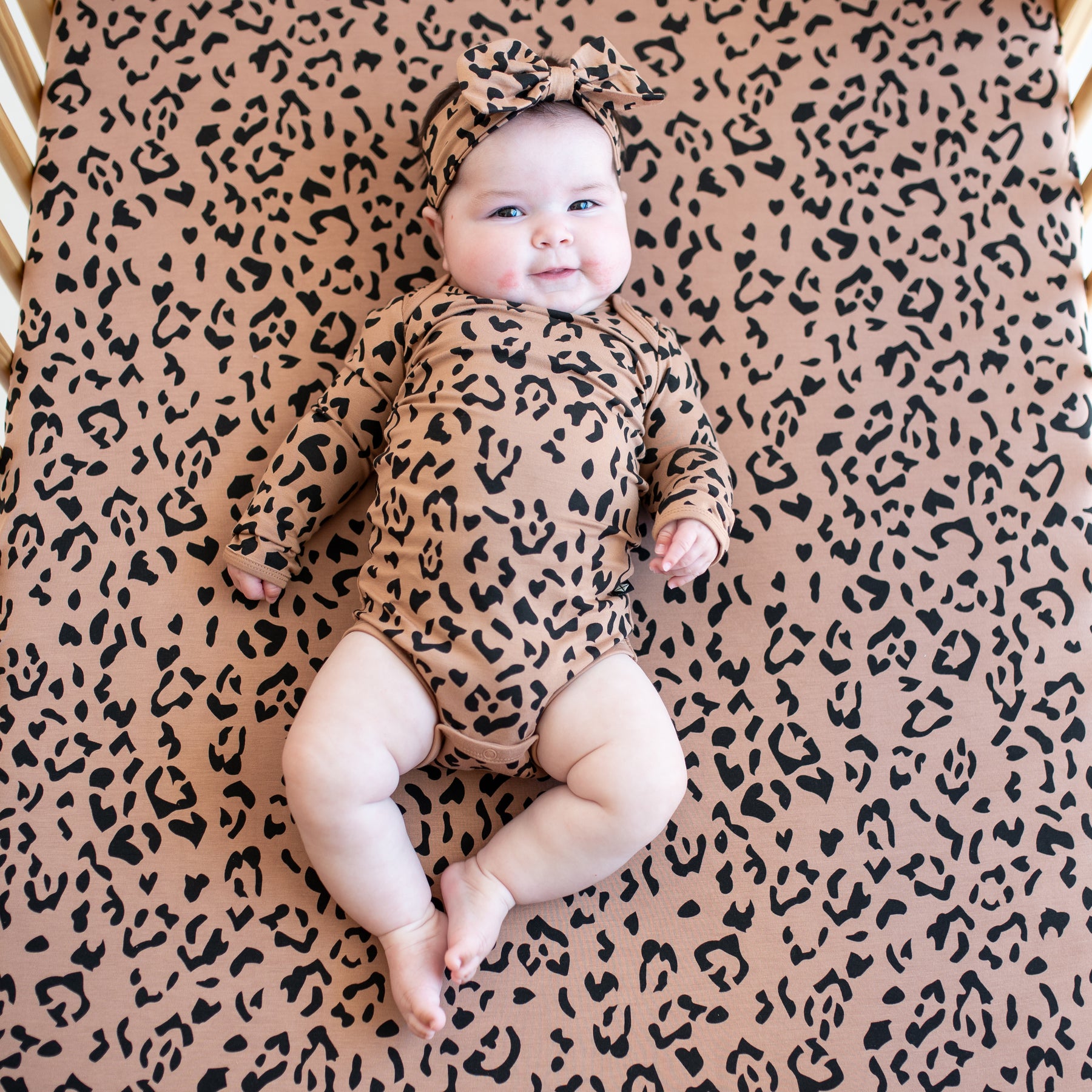 Infant laying on a Crib Sheet in Latte Leopard wearing the matching long sleeve bodysuit and bow headband
